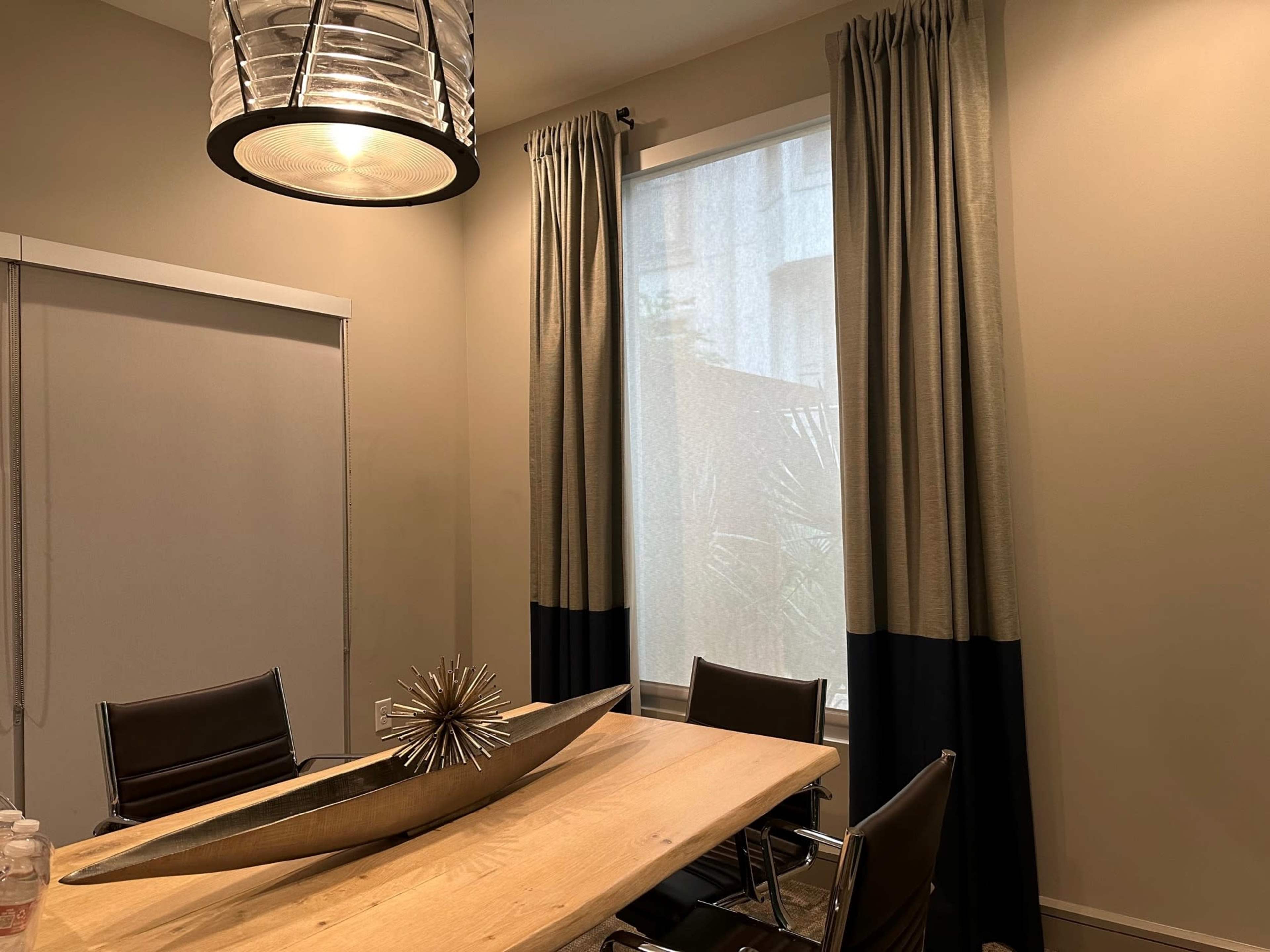 A modern dining area features a wooden table with a decorative centerpiece, surrounded by black leather chairs and dressed with long curtains on a window.