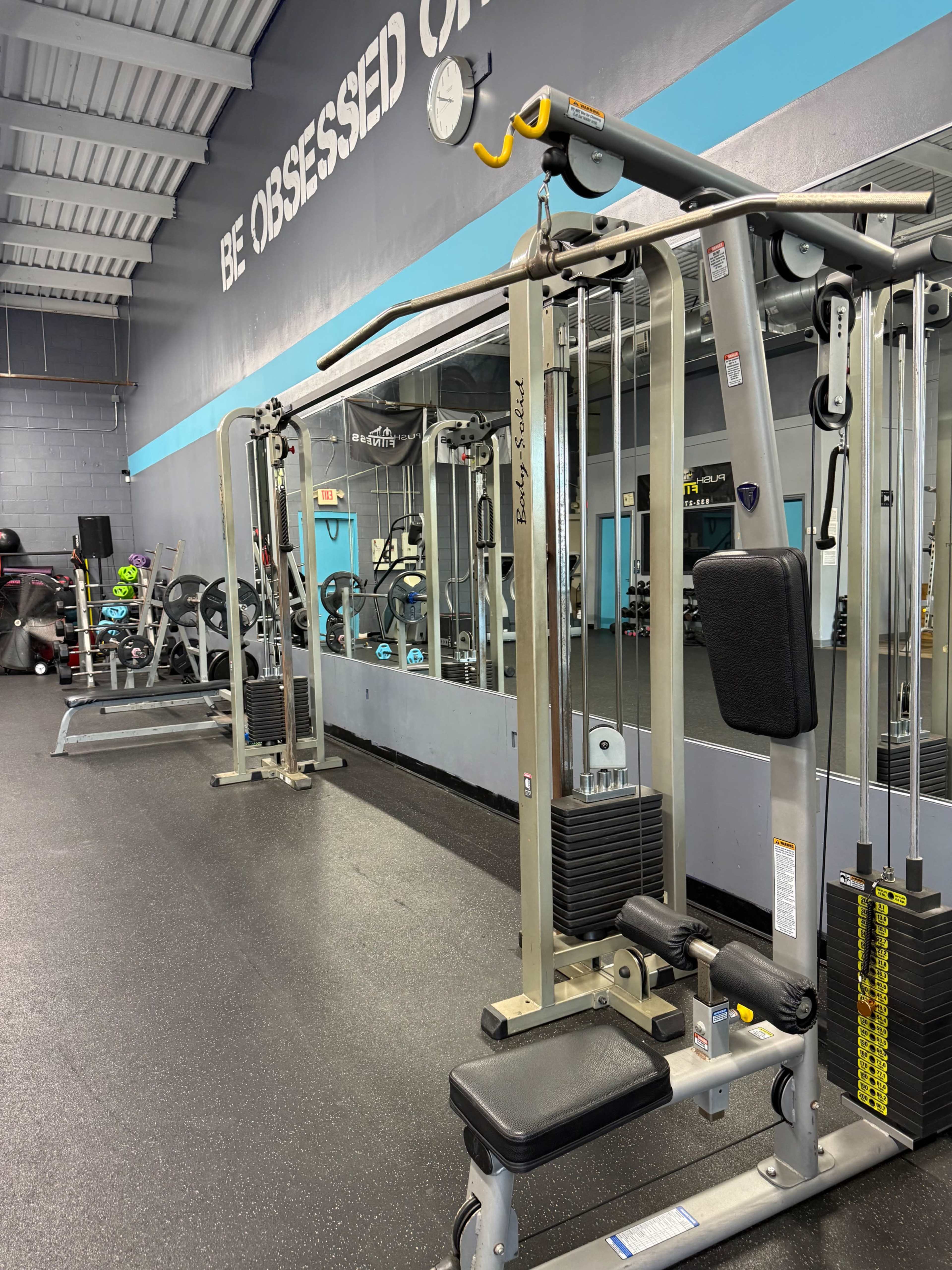 The image shows a fitness gym interior with a cable machine in the foreground and exercise equipment visible in the background, along with a large mirror on the wall.