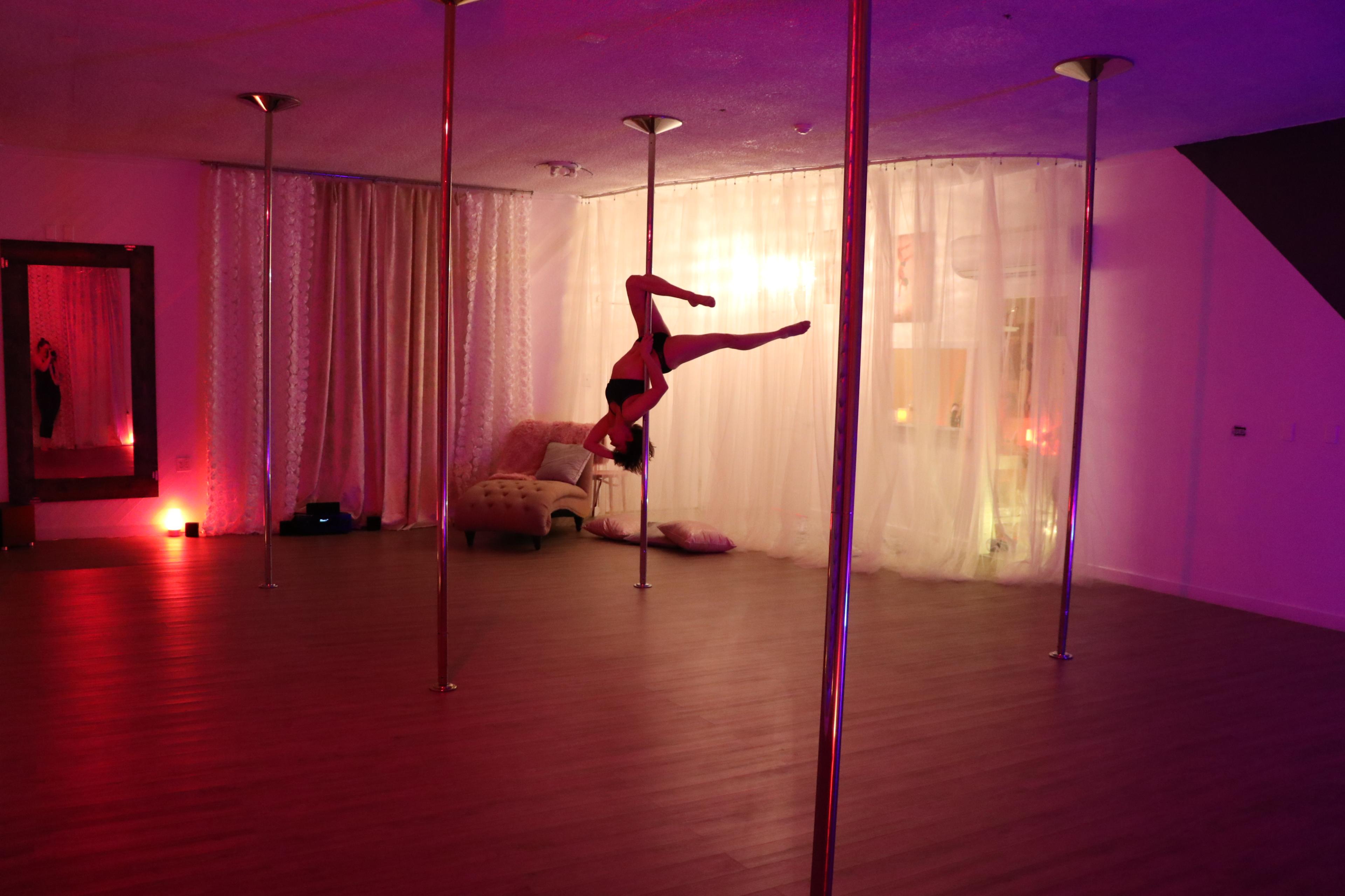 A dancer performs an inverted pose on a pole in a dimly lit studio with soft pink lighting and sheer curtains.