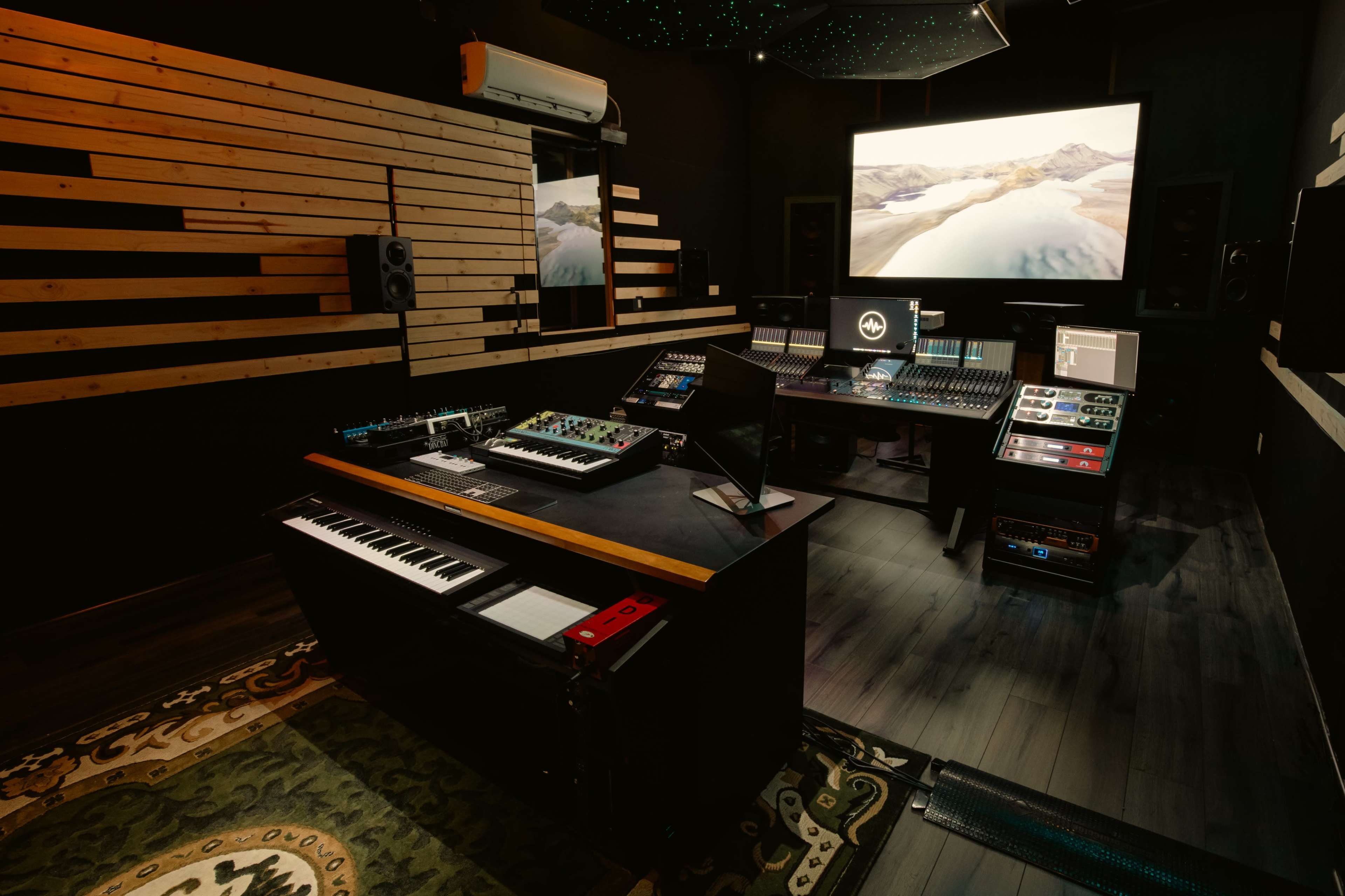 A modern recording studio with various music production equipment, including a digital mixing console, keyboards, and a large screen displaying a landscape.