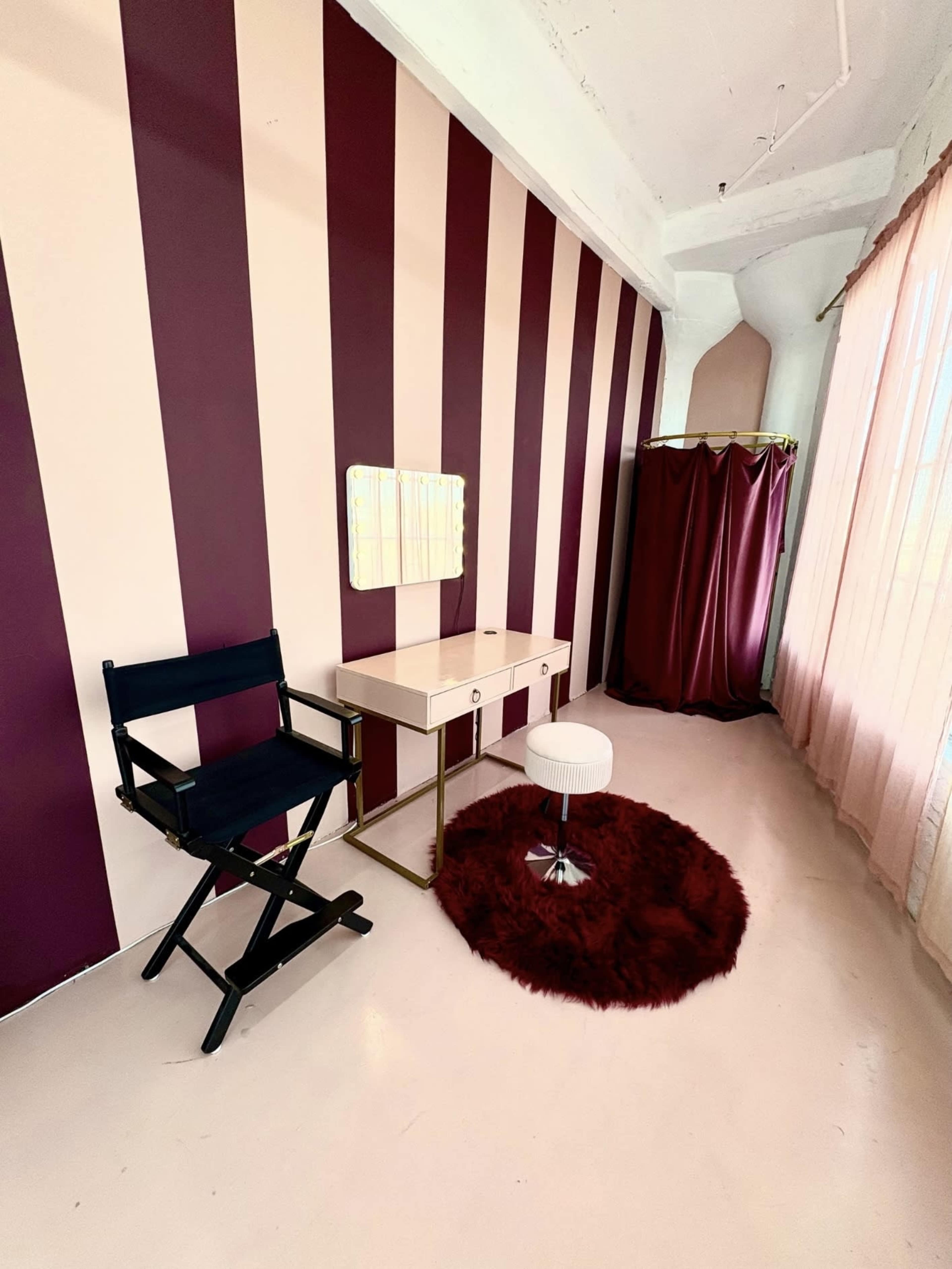 The image shows a room with vertical striped walls in pink and burgundy, featuring a vanity table with a mirror, a black director's chair, a white stool, and a red circular rug.