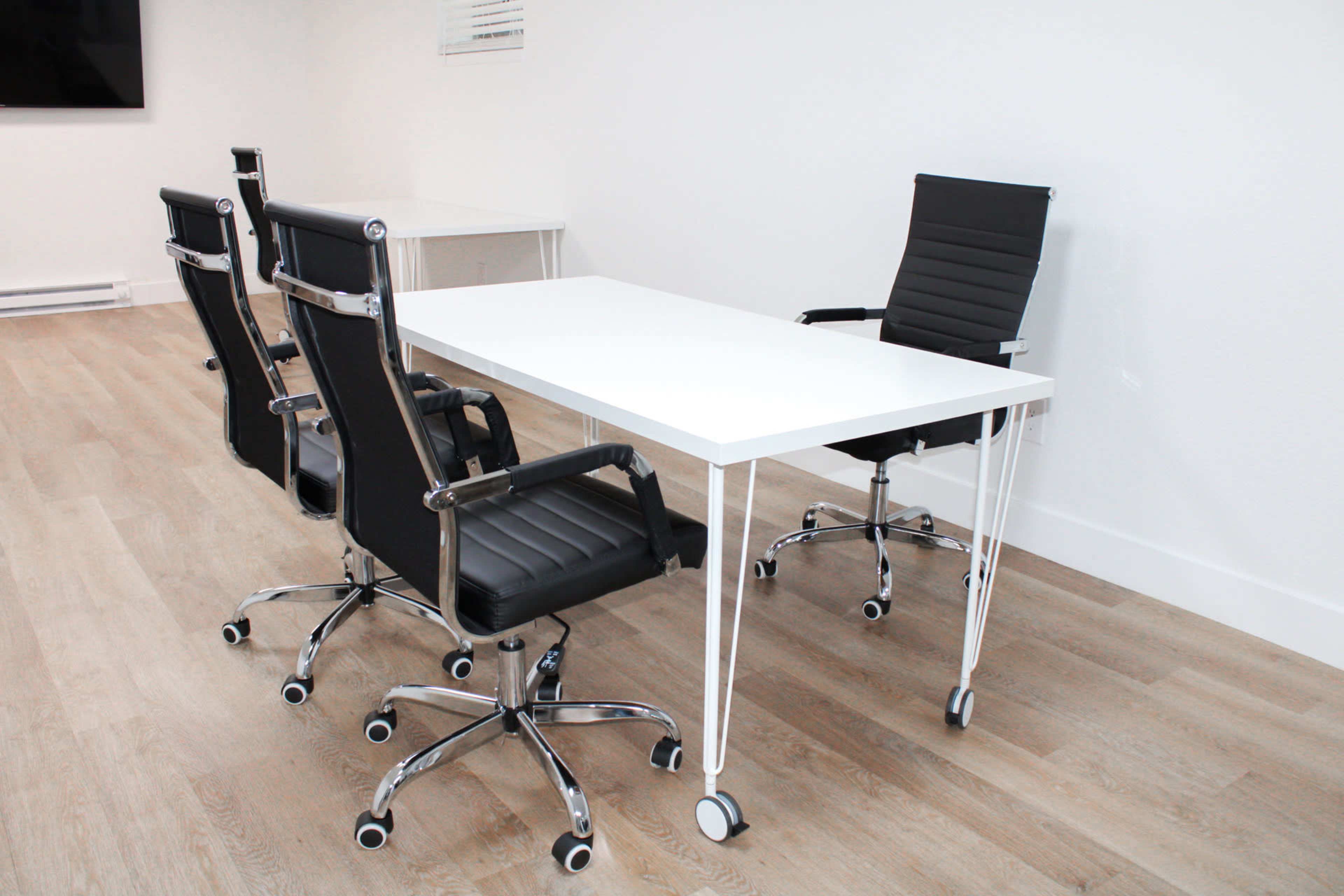 A modern conference room features a white table surrounded by four black office chairs on a light wood floor.