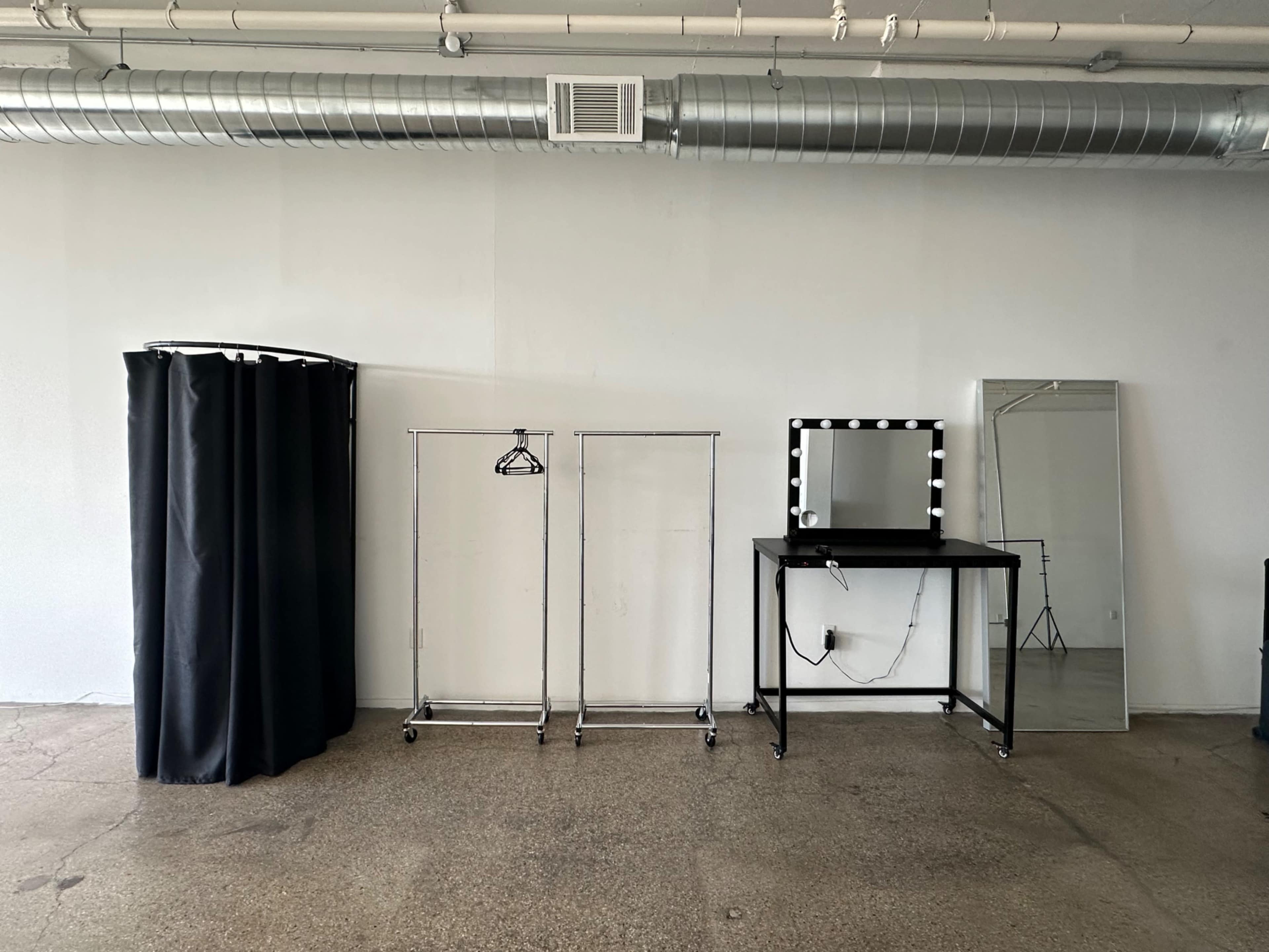 The image shows a minimalist dressing area featuring a black curtain, two clothing racks on wheels, a lit makeup vanity with a mirror, and a standing mirror against a plain wall.