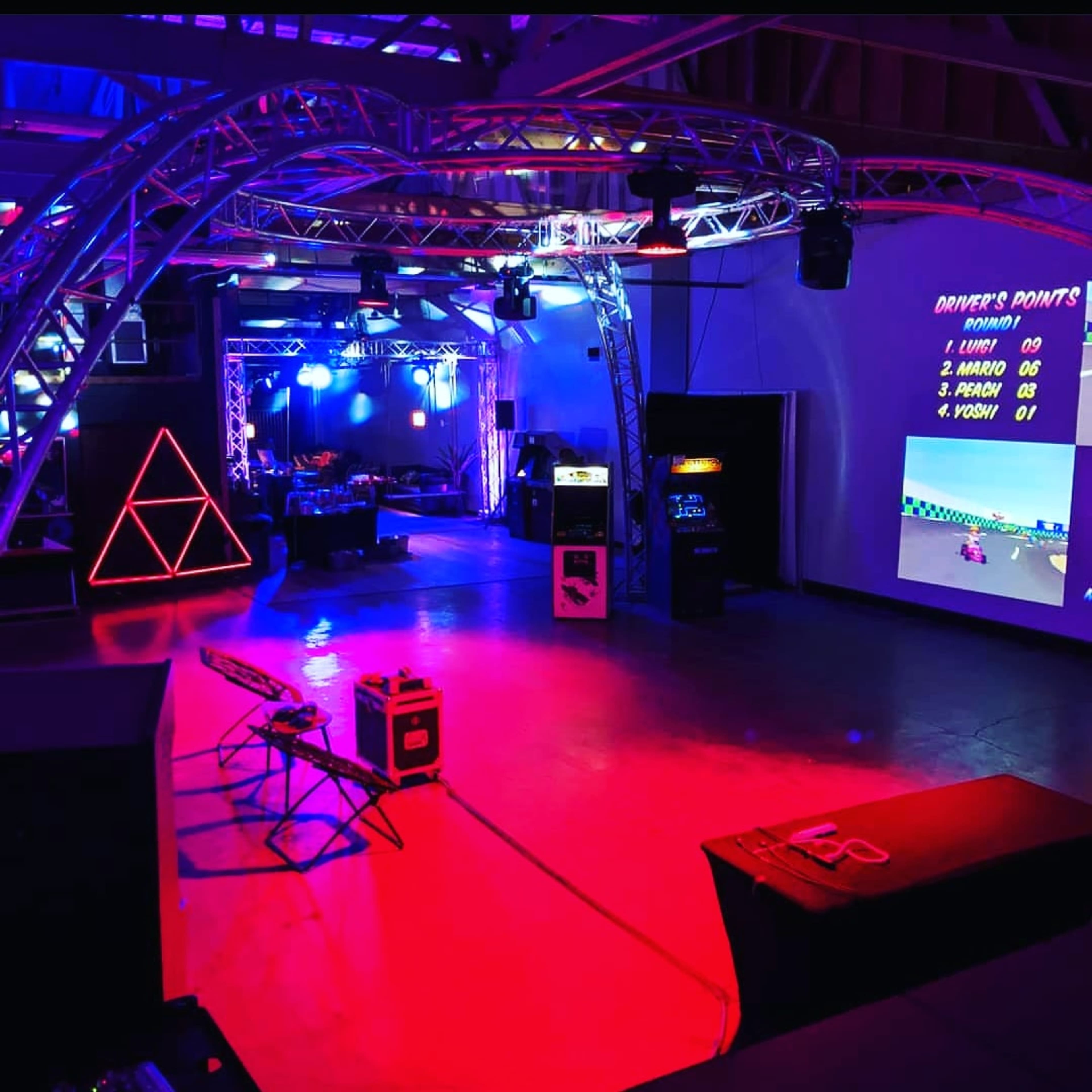 A brightly lit arcade space with a racing video game displayed on a screen, surrounded by various arcade machines and colorful LED lighting.