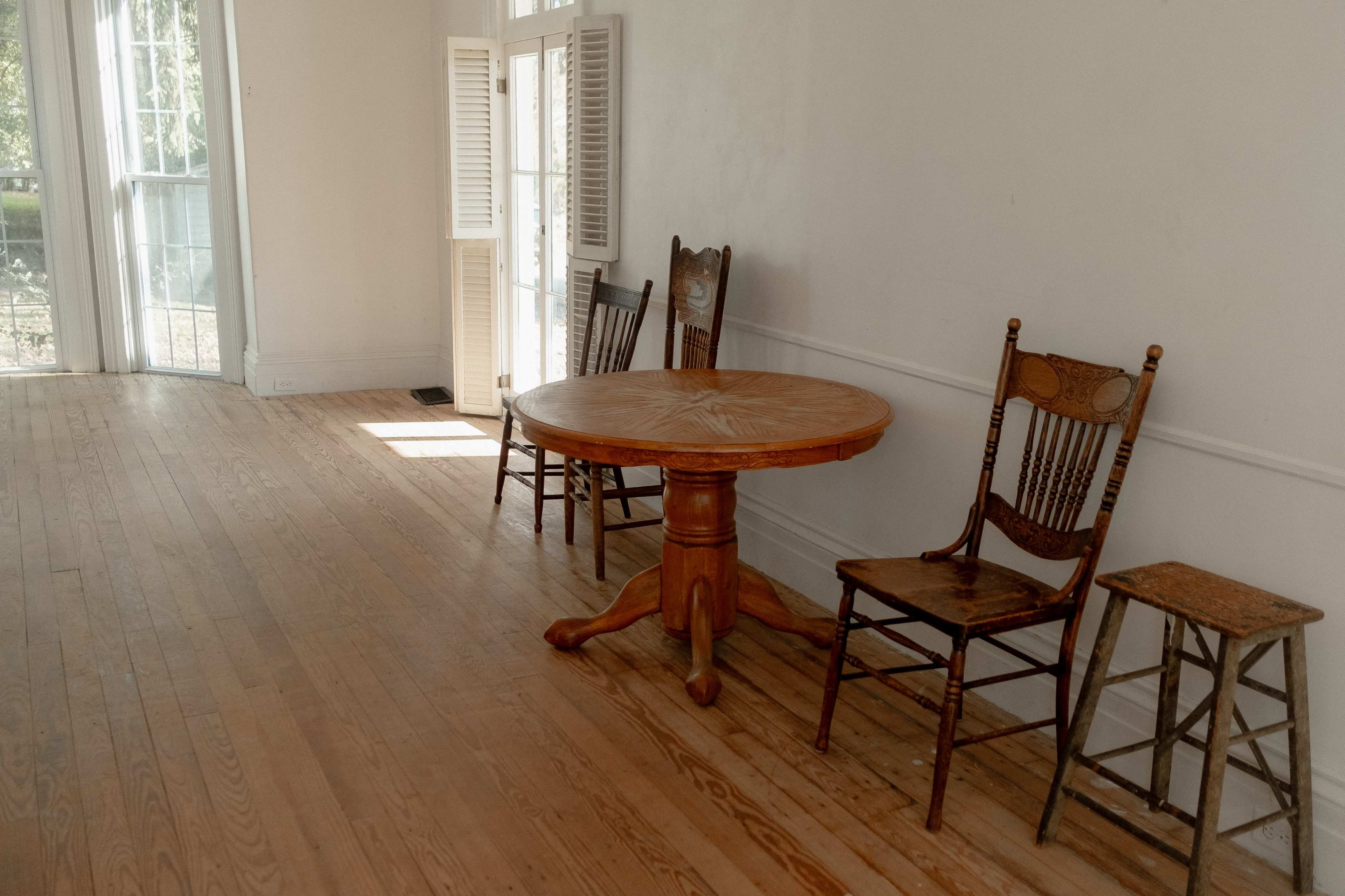 A round wooden table is positioned in a light-filled room alongside three wooden chairs on a polished wooden floor.