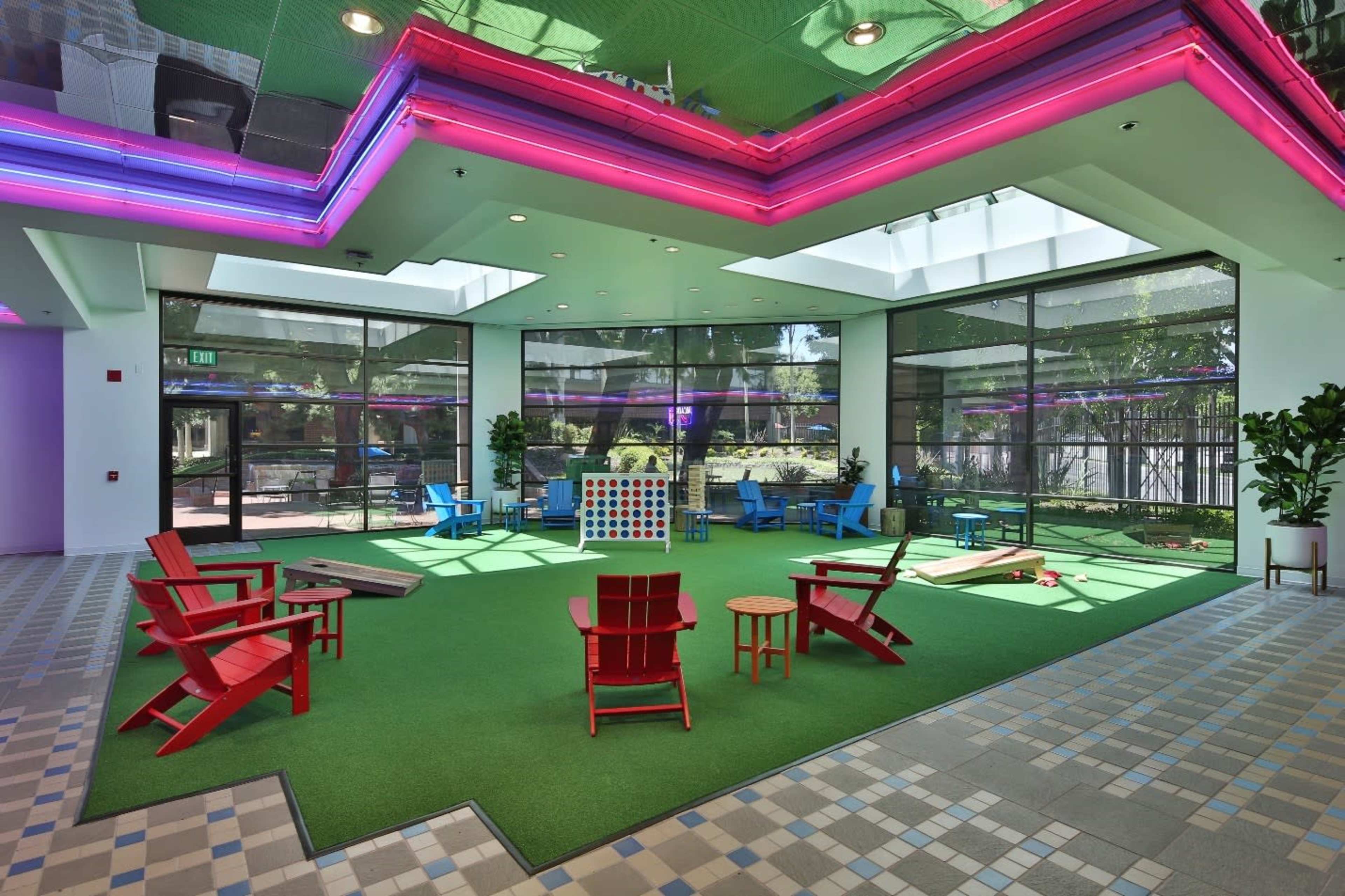 An indoor recreation area featuring colorful chairs, a large Connect Four game, and green carpeting with natural light streaming in through large windows.
