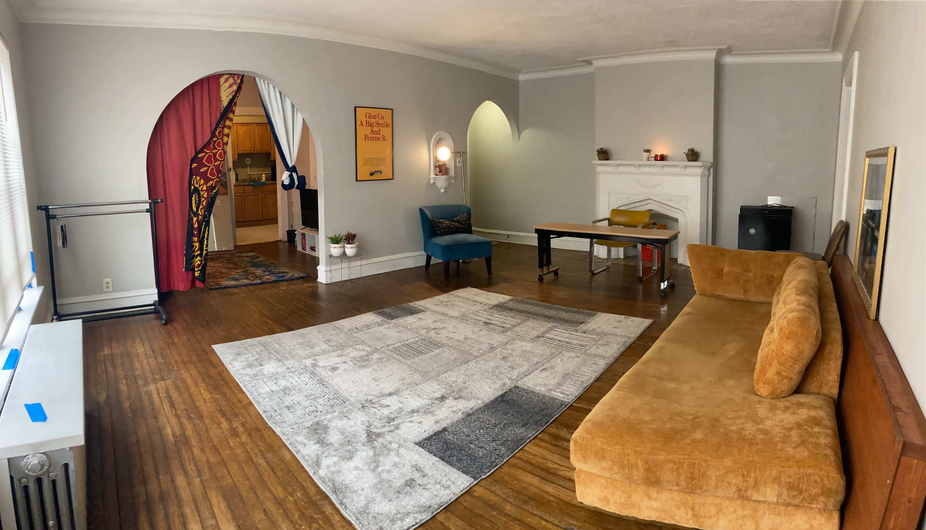 A spacious room with a wooden floor, a large area rug, a beige couch, a table with a chair, and an archway leading to another area with a kitchenette.