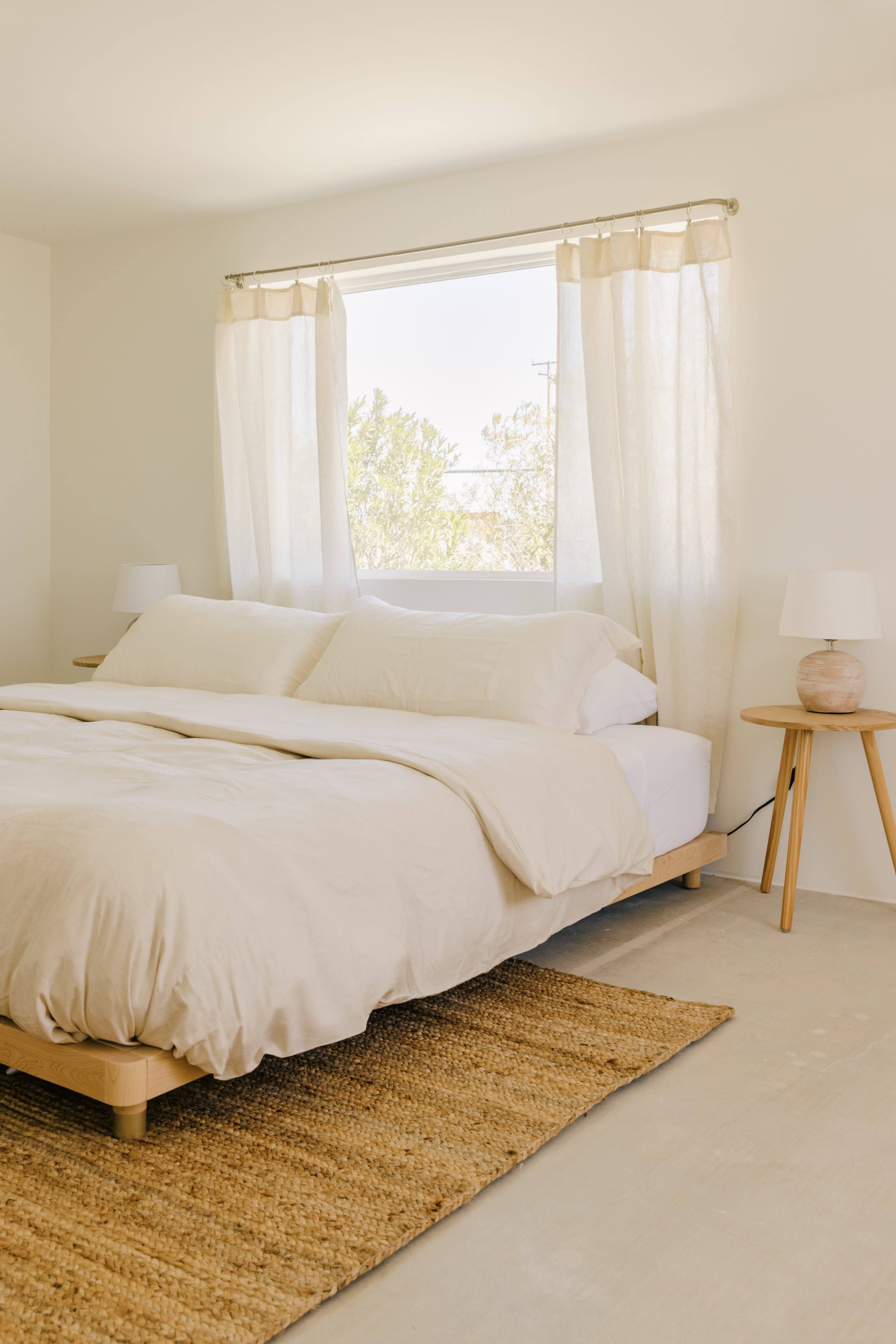 The image shows a simple bedroom featuring a bed with a light-colored duvet, two bedside lamps, and a woven rug on a concrete floor.