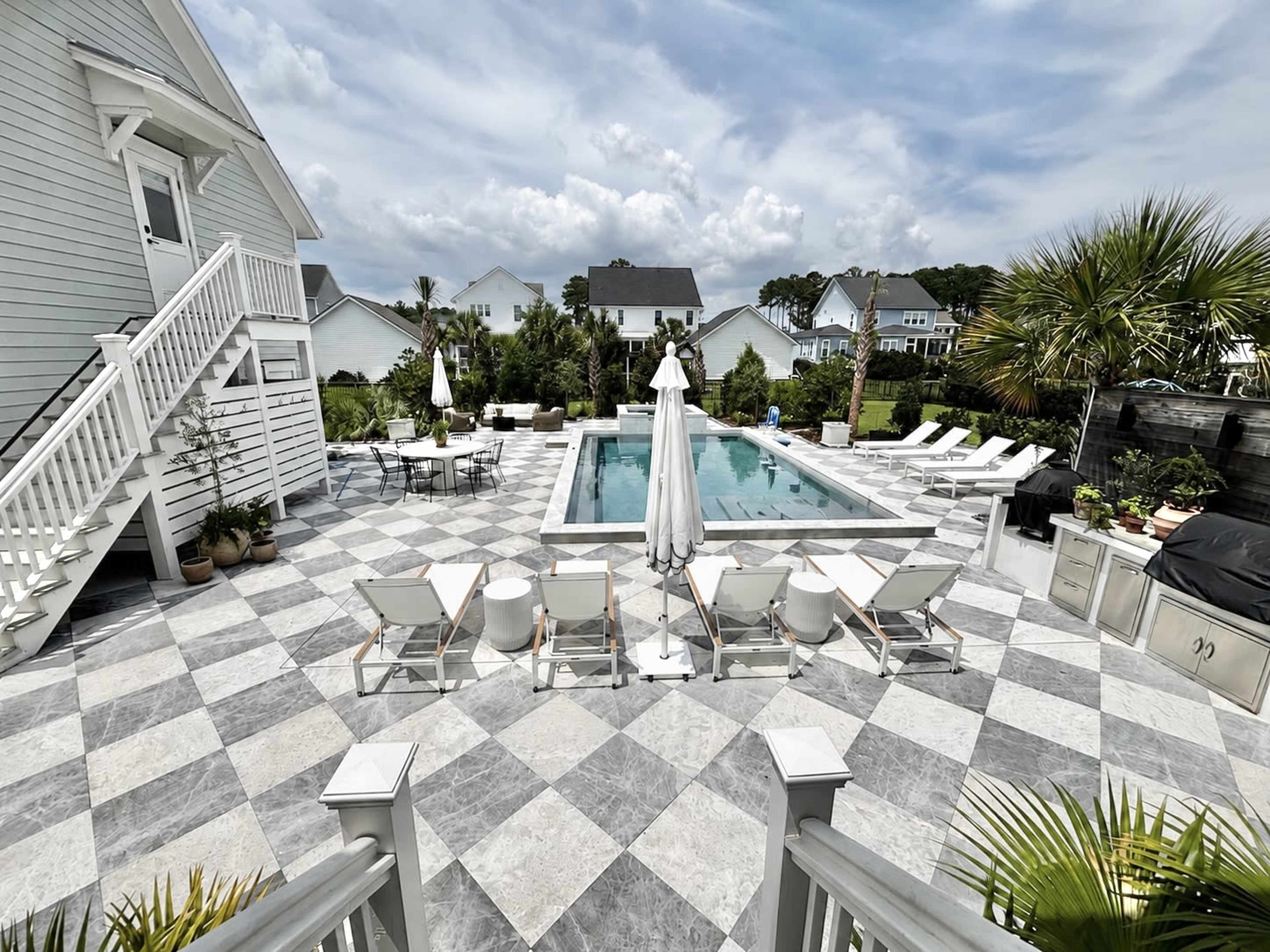 A backyard with a rectangular swimming pool surrounded by lounge chairs, patio furniture, and a white umbrella, set against a backdrop of houses and palm trees.