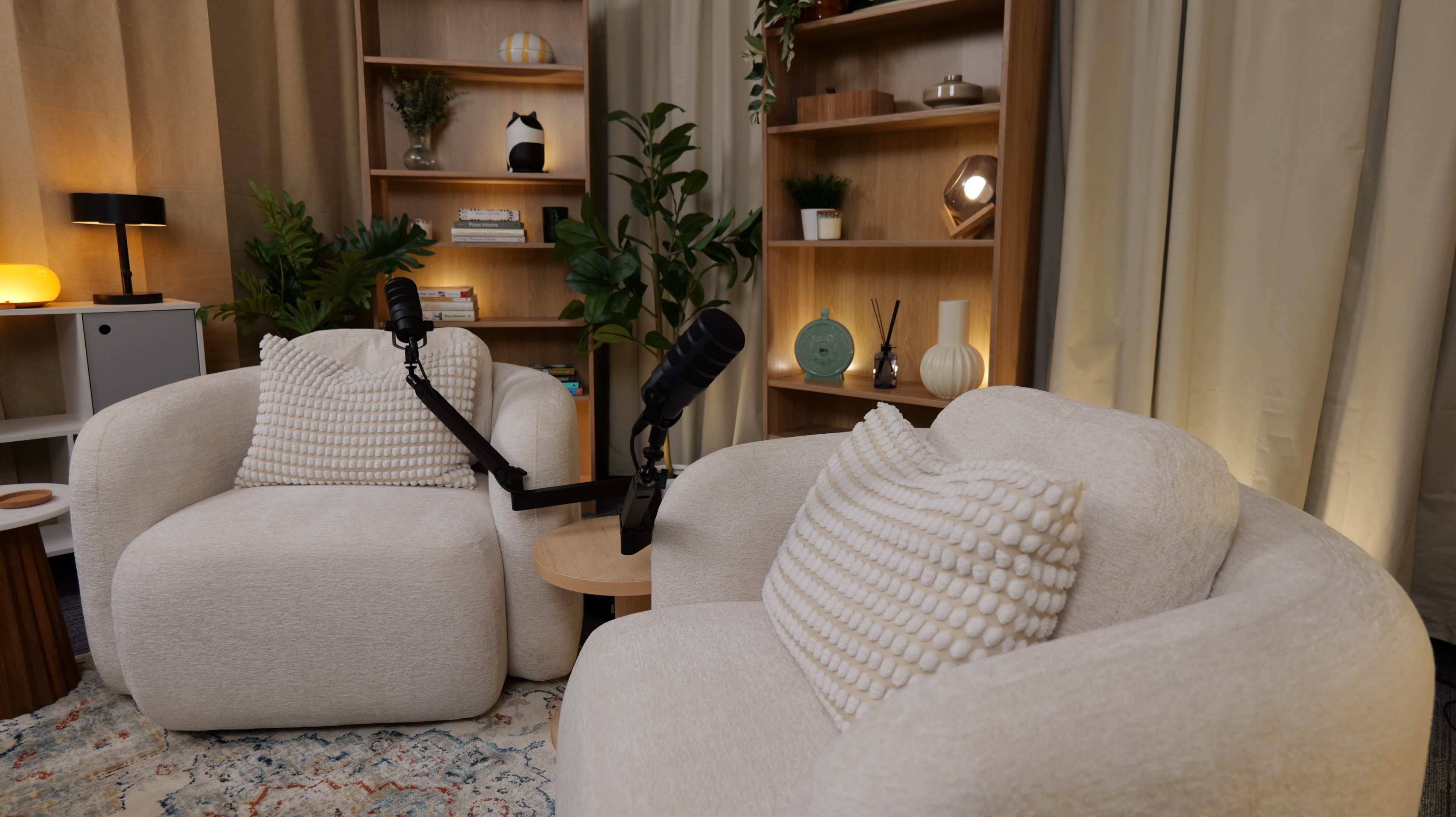 The image shows two comfortable, upholstered chairs with a microphone setup between them, surrounded by shelves adorned with decorative items and plants.