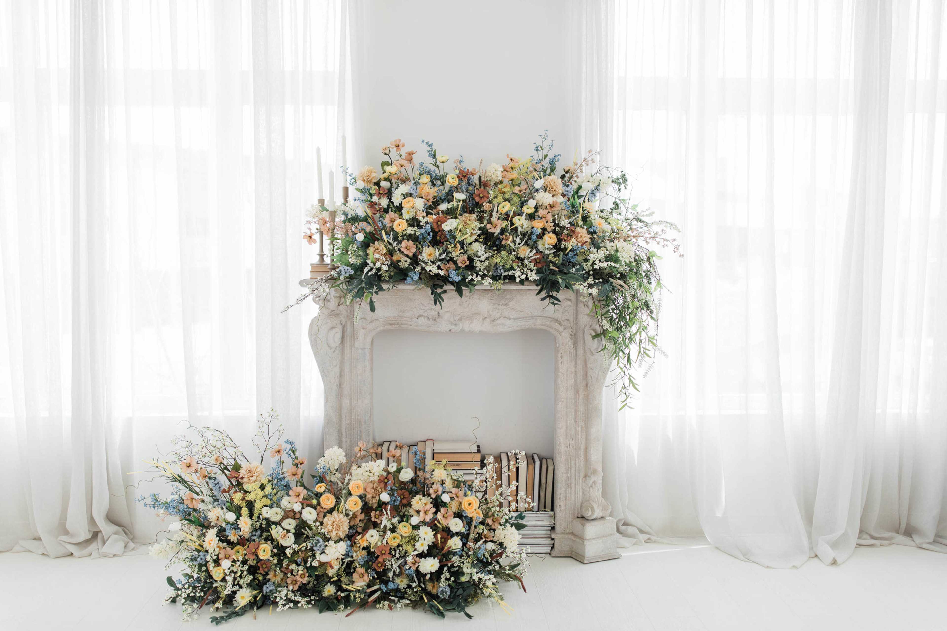 A floral arrangement adorns a white fireplace, complemented by stacks of books, against a backdrop of sheer curtains.
