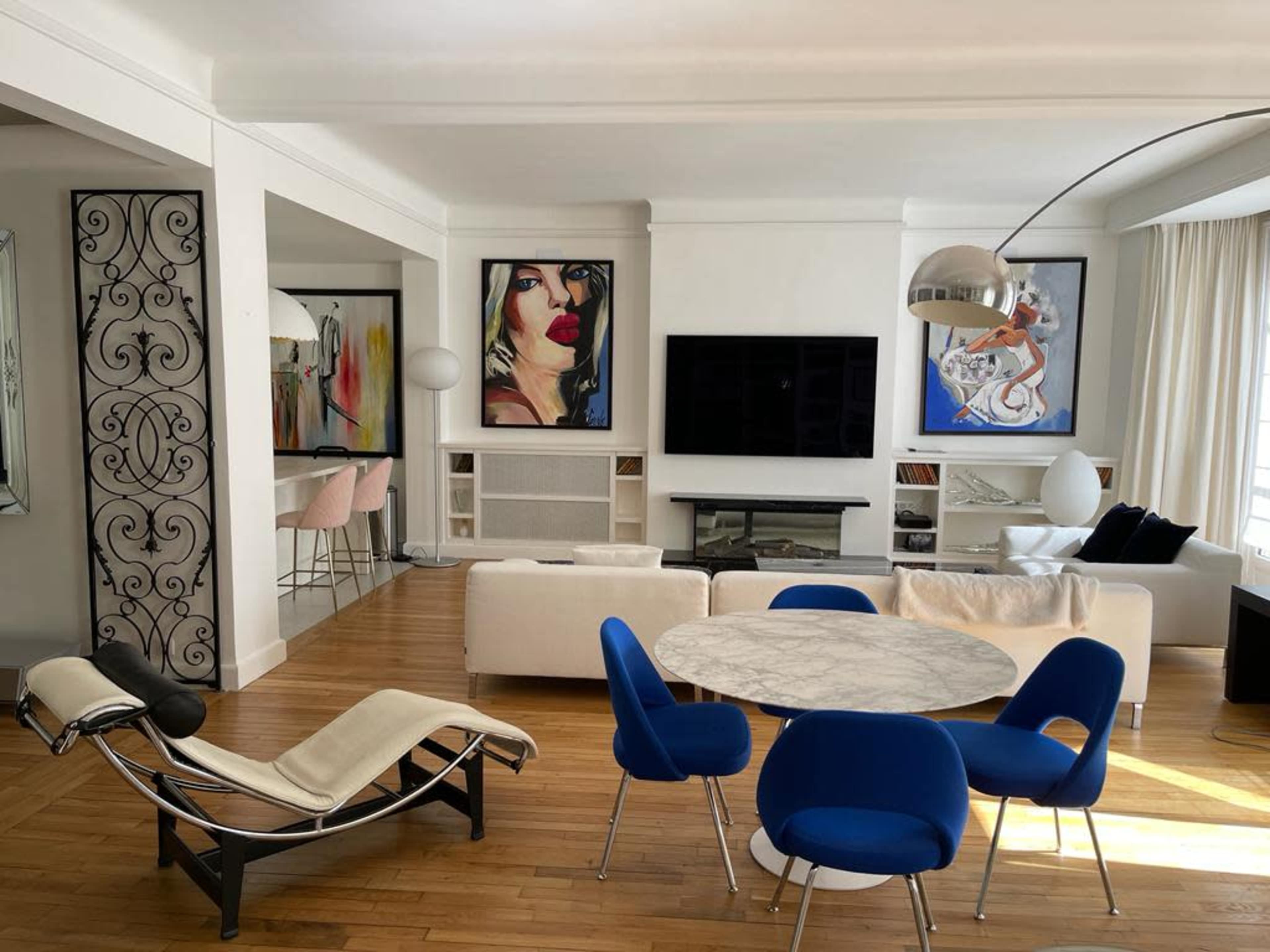 A modern living space features a marble dining table surrounded by blue chairs, a white sofa, and artwork on the walls, with a large TV and a fireplace in the background.