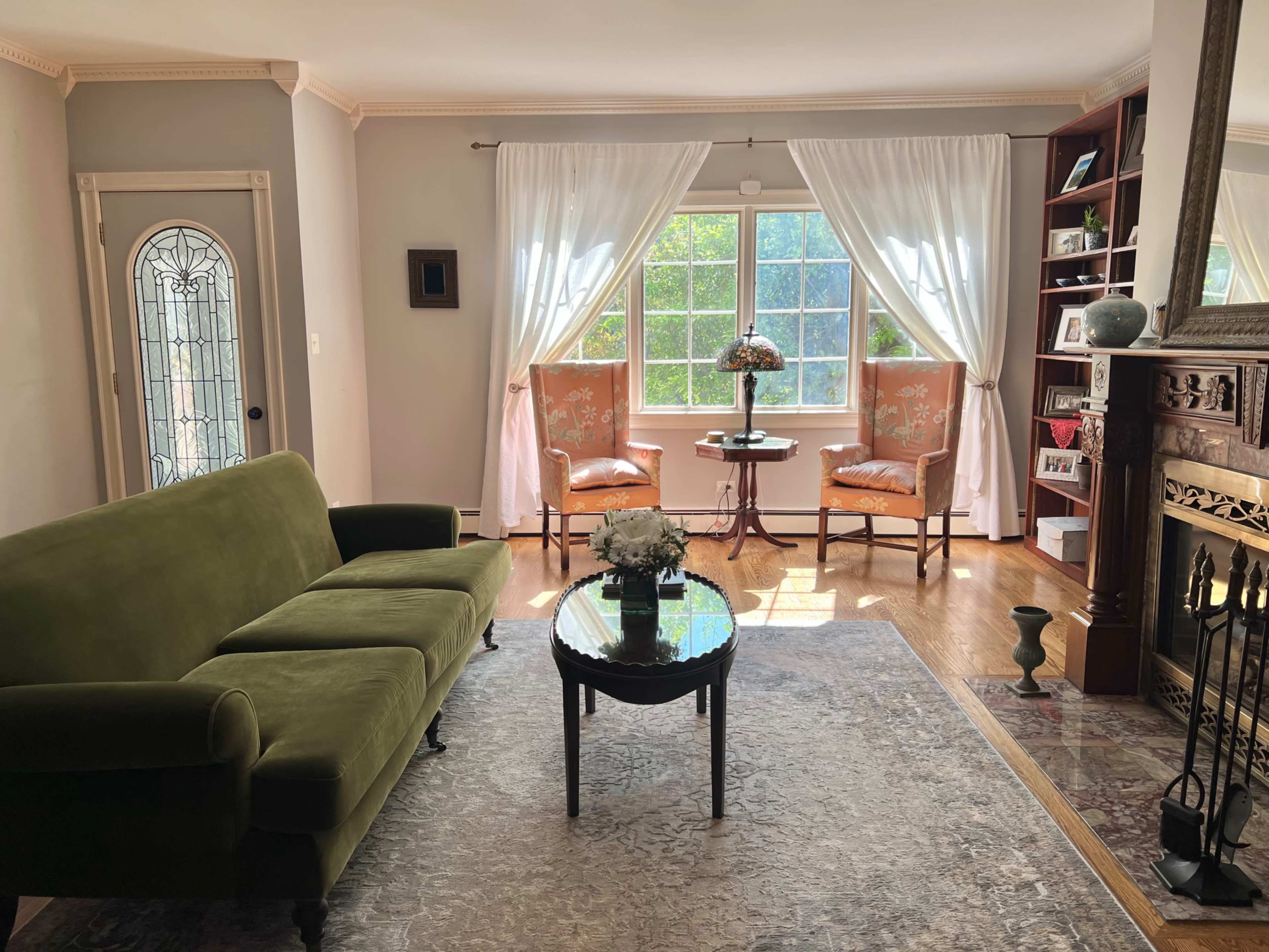 The image shows a living room with a green sofa, a round coffee table, two chairs by a window, and a fireplace on one side.
