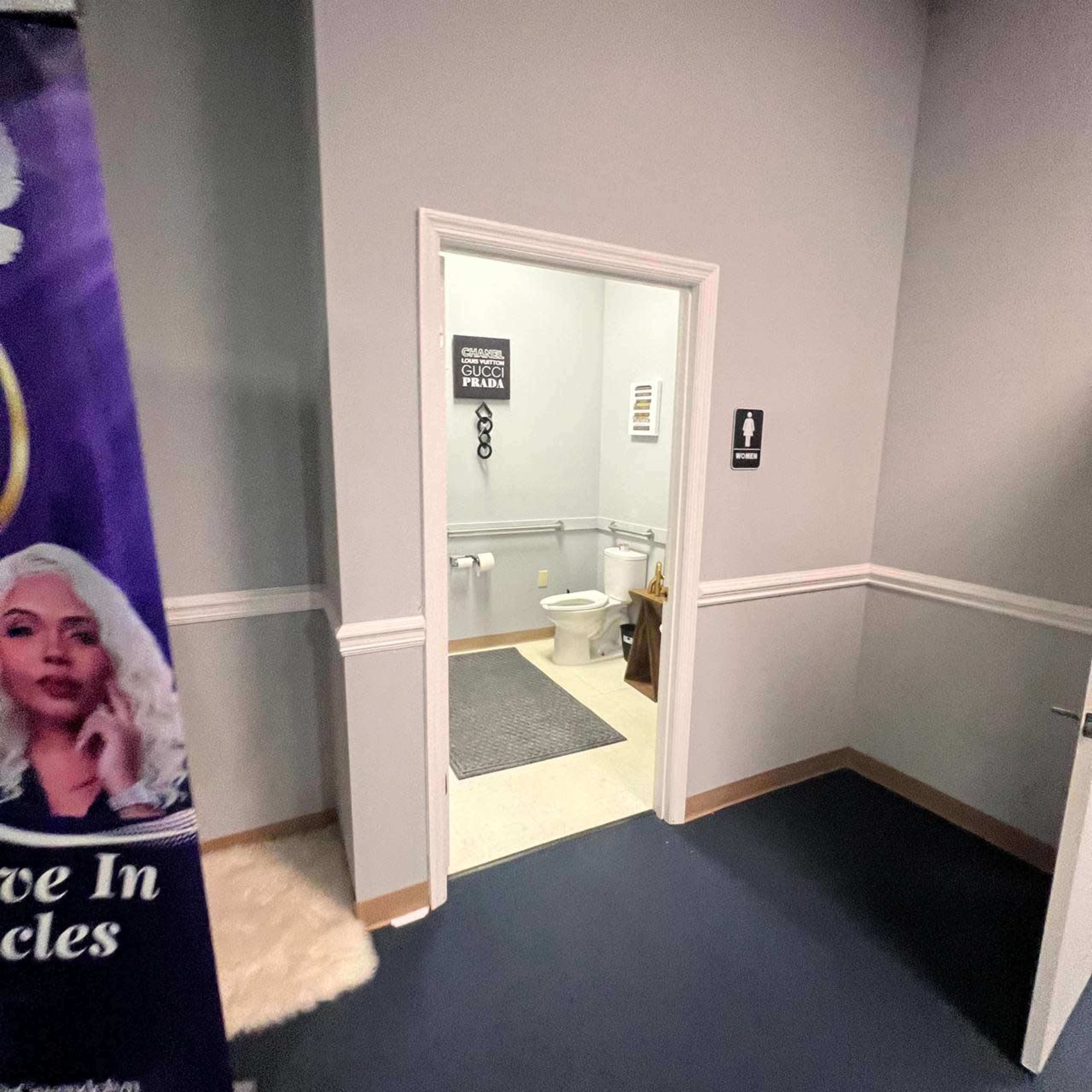 The image shows an entrance to a restroom with a toilet visible inside, flanked by light gray walls and a decorative sign.