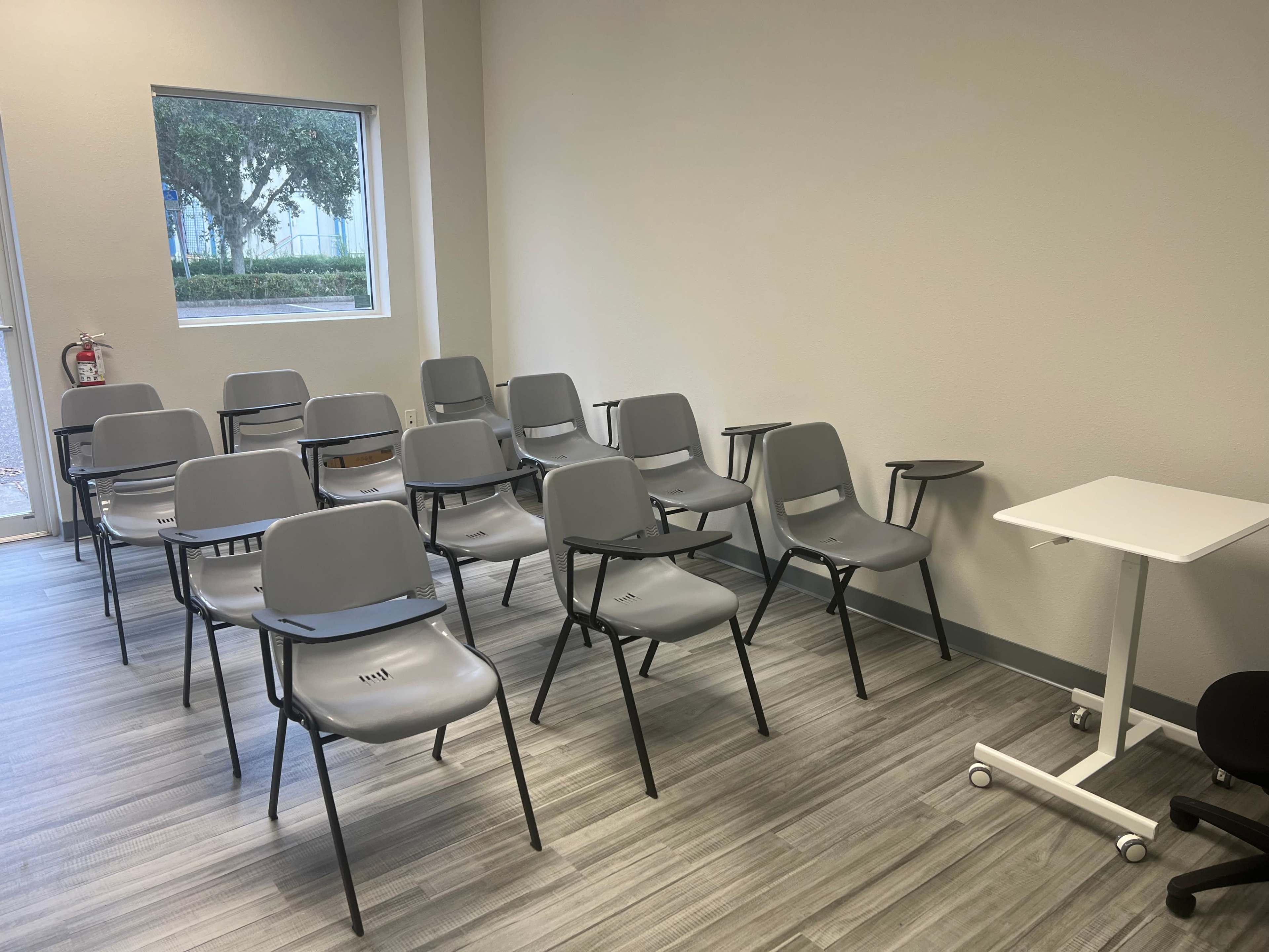 The image shows a sparsely furnished classroom with several gray chairs arranged in rows and a small table in the corner.