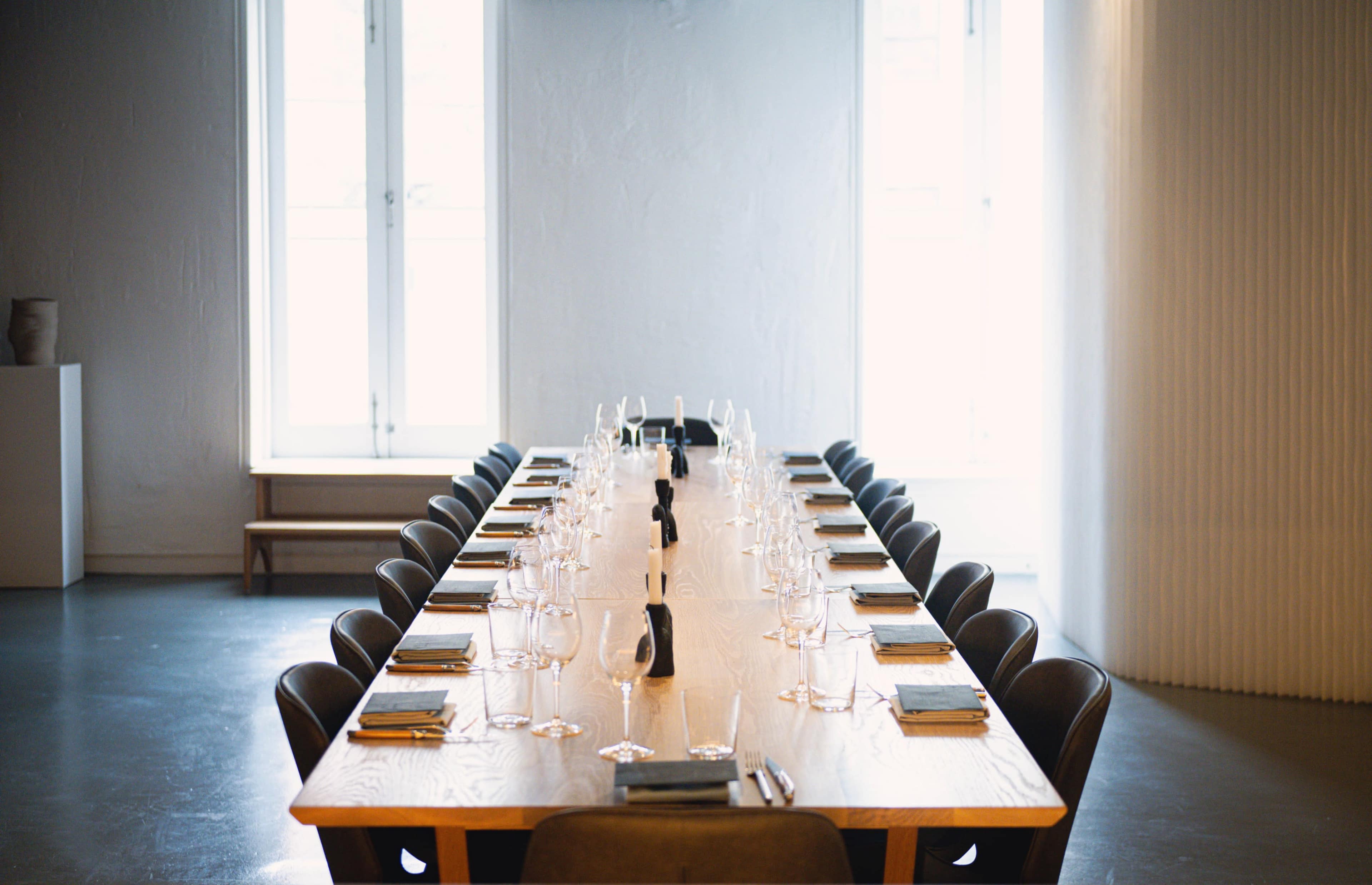 A long wooden dining table is set with plates, utensils, and glasses, positioned under large windows in a minimalist interior space.