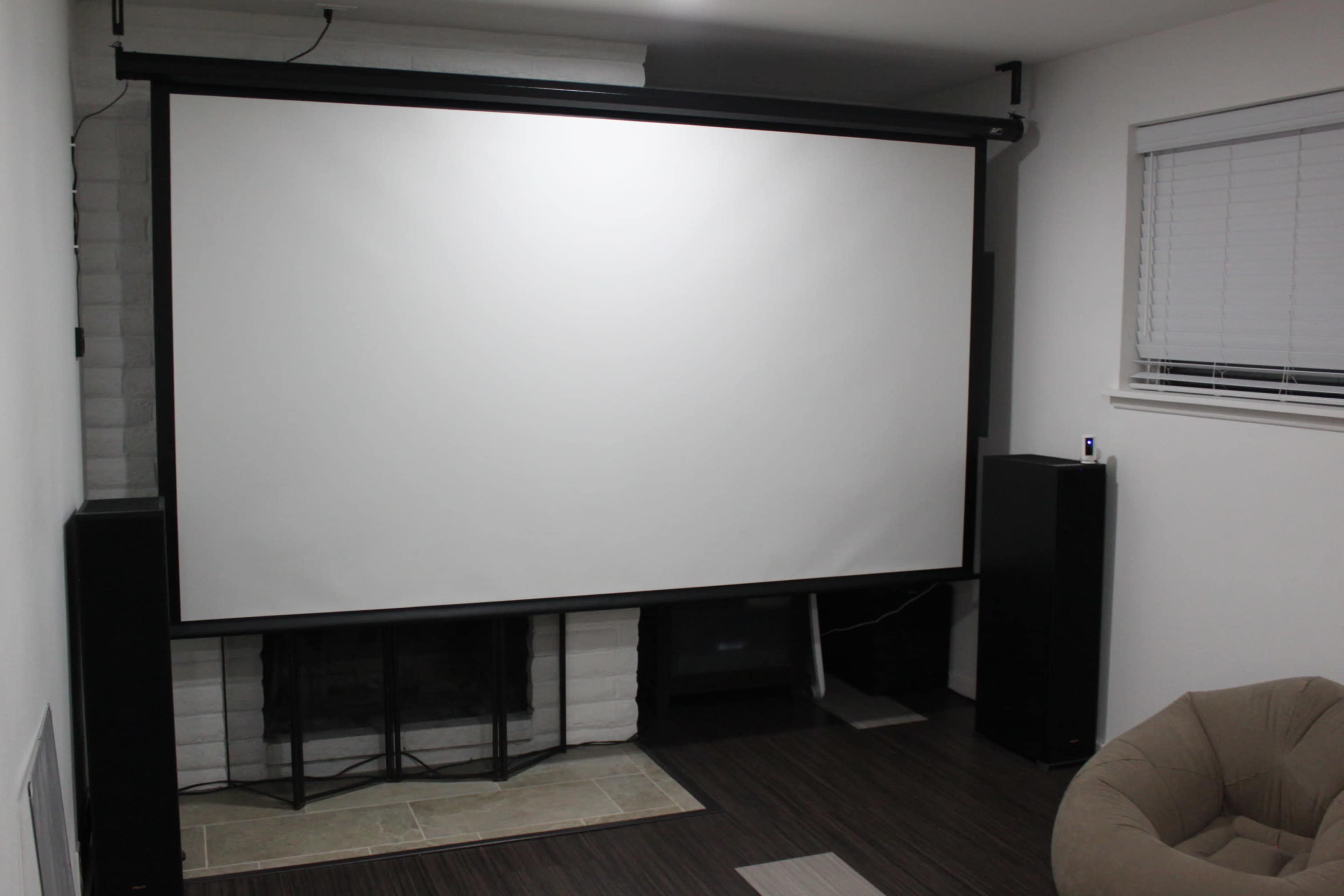 The image shows a blank projection screen mounted on a wall in a room with a fireplace and a light-colored floor.