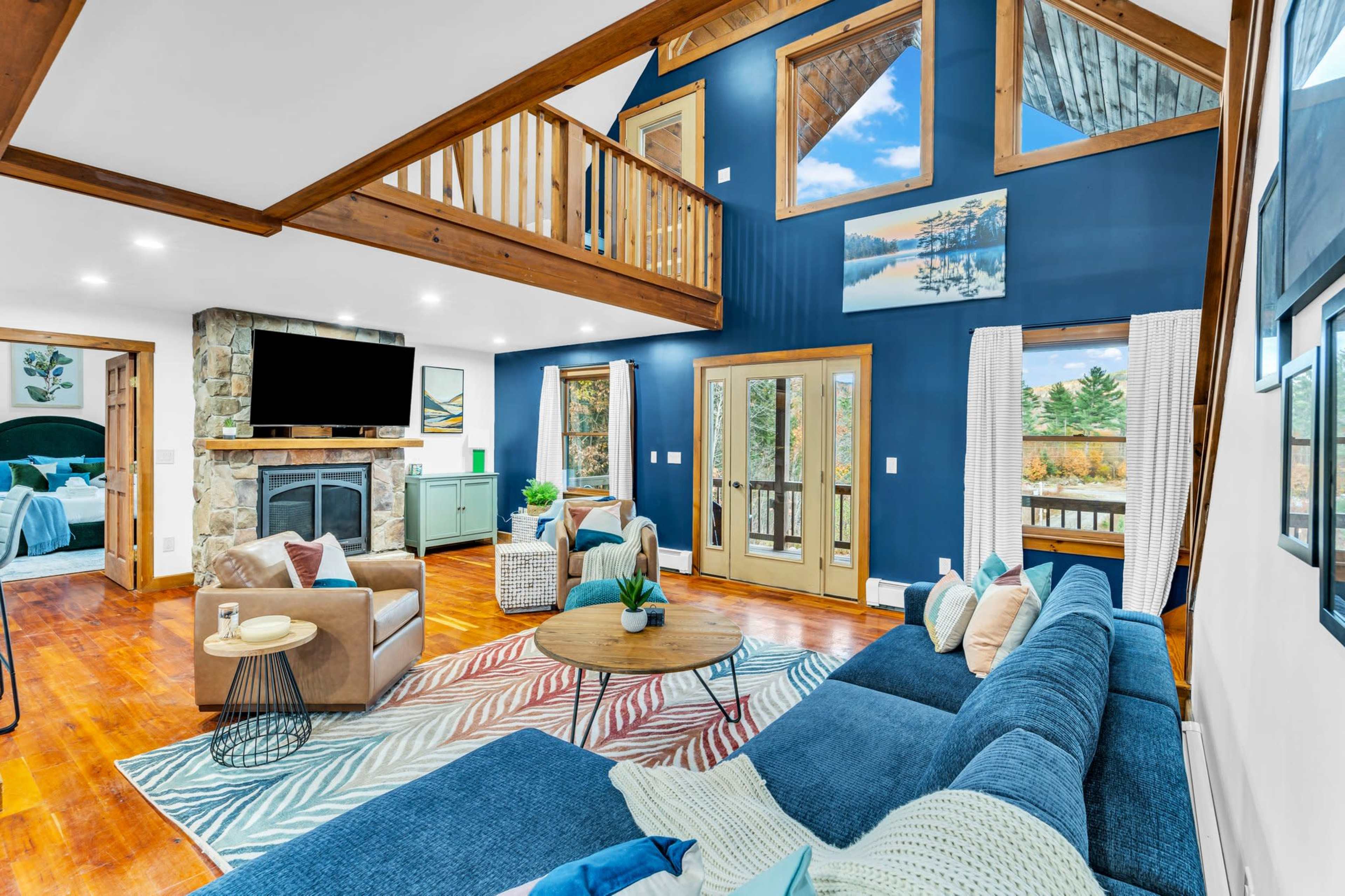 A modern living room with a blue accent wall, large windows, a stone fireplace, and a sectional sofa arranged around a coffee table.