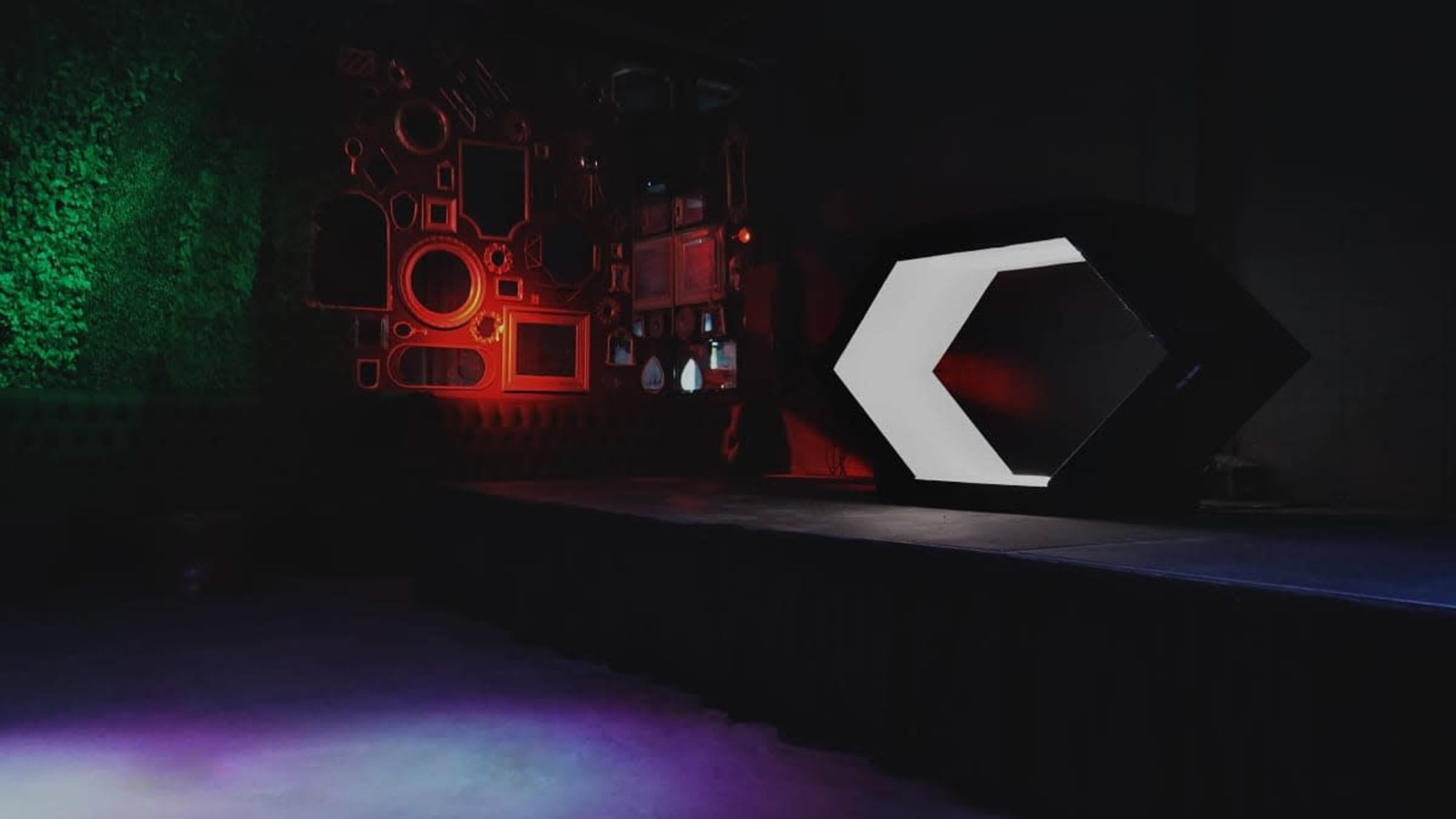 The image shows a stage with a geometric, hexagonal structure illuminated in white and red lights, set against a textured wall with decorative mirrors.