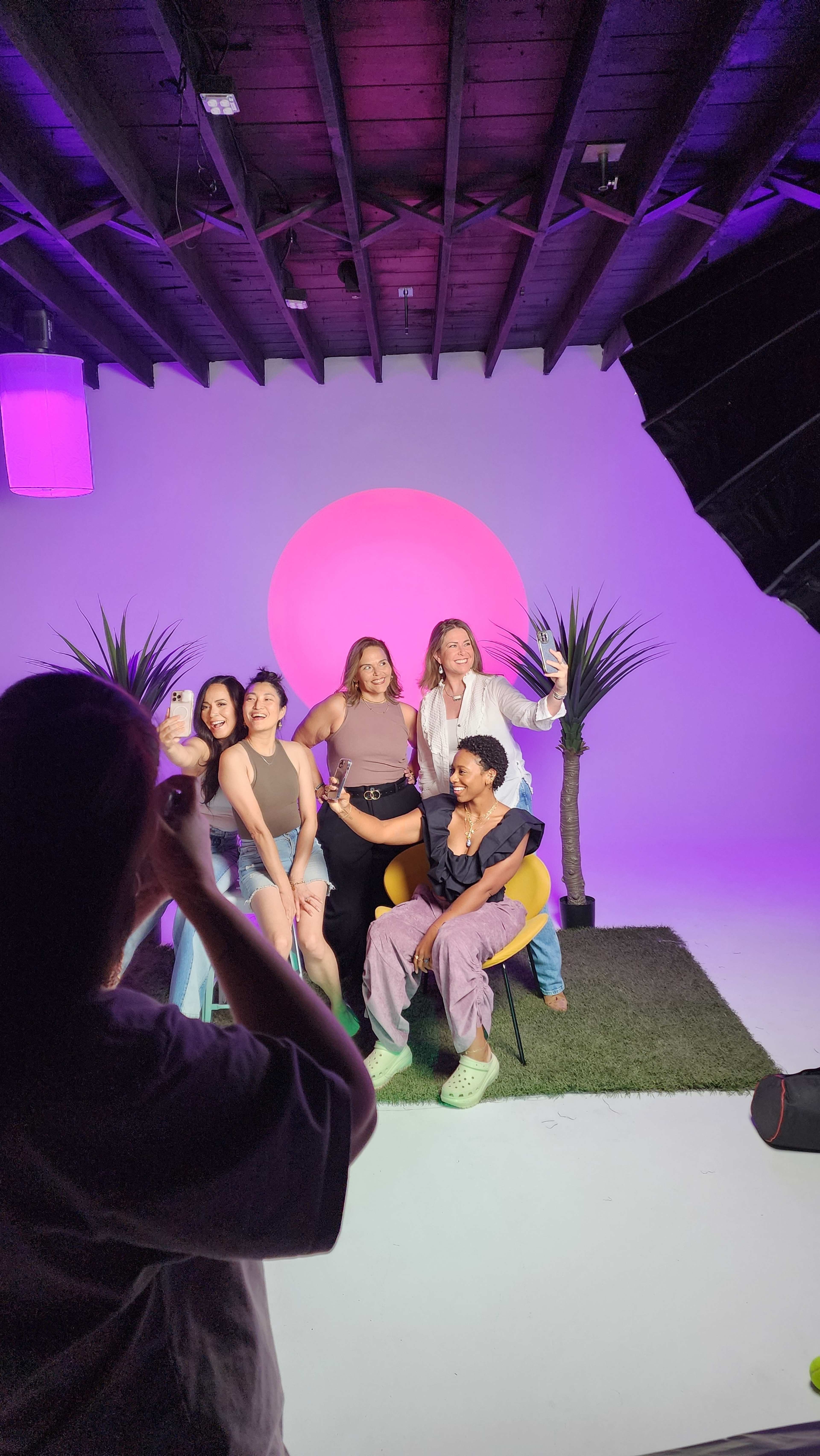 A group of five people poses for a photo in a studio with a pink backdrop and decorative plants.