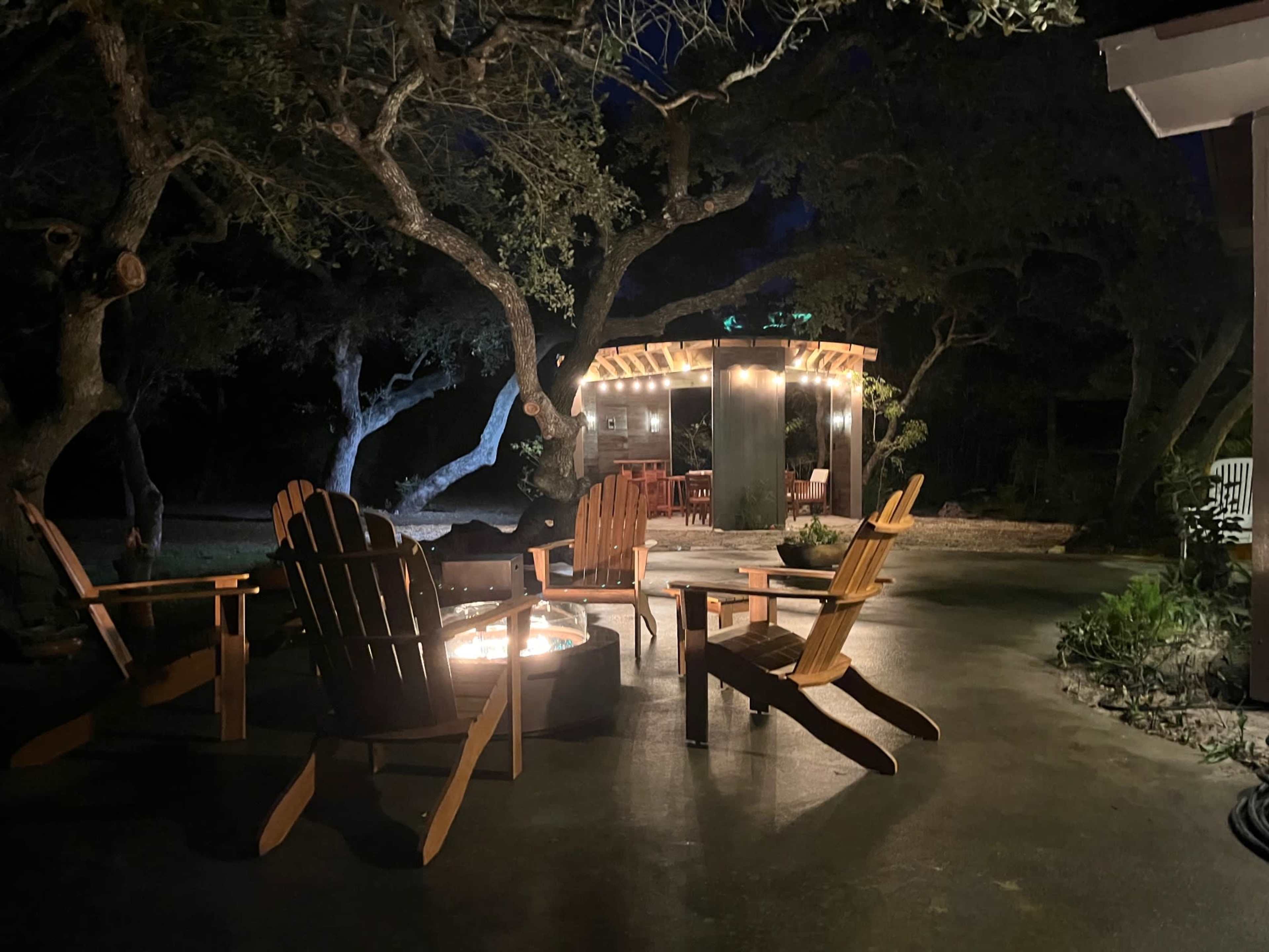 A circle of wooden chairs surrounds a fire pit in a dimly lit outdoor space featuring a gazebo adorned with string lights.