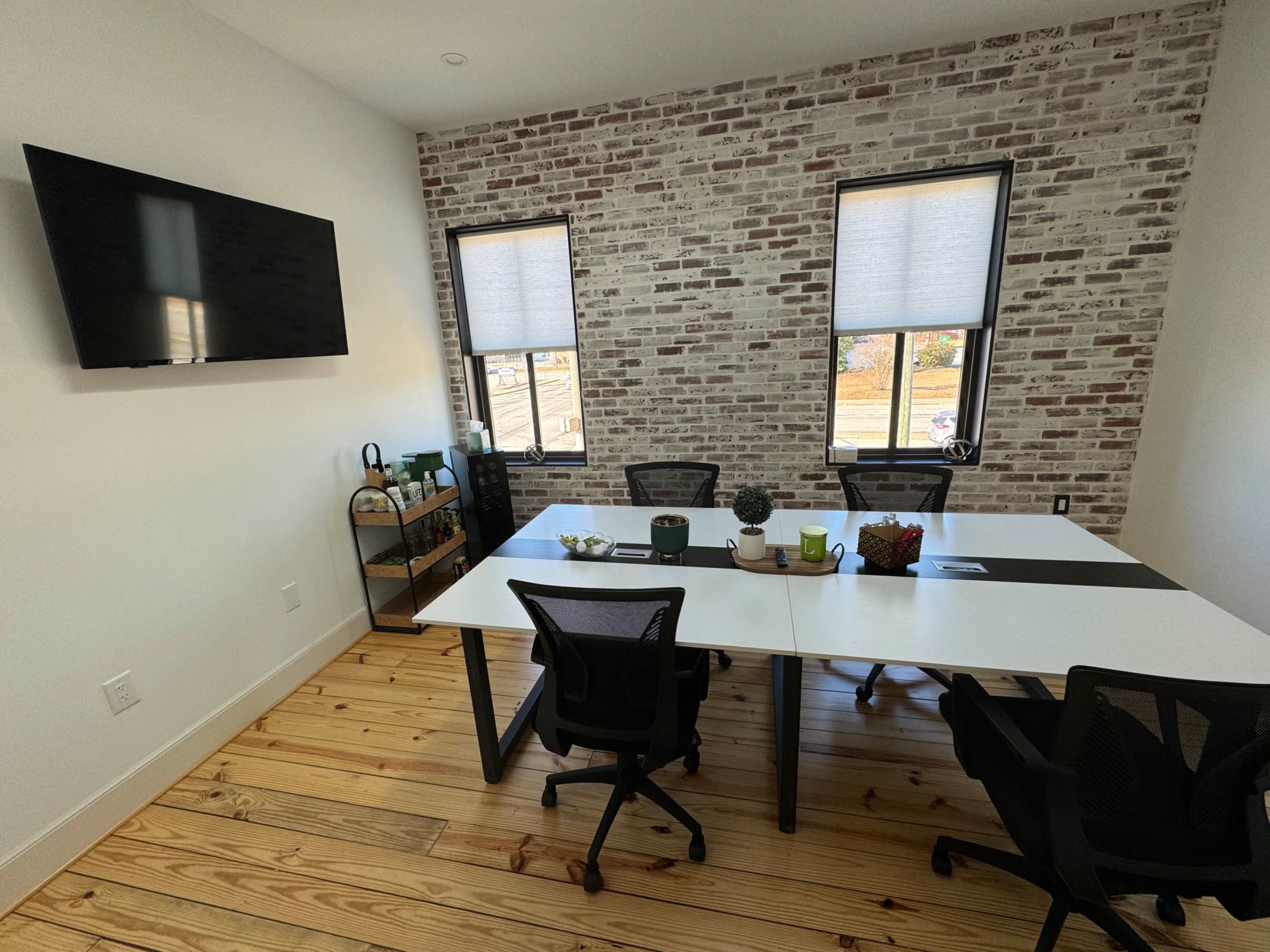 A modern meeting room featuring a long table with black chairs, a television mounted on the wall, and large windows with blinds, set against a brick wall and wooden flooring.