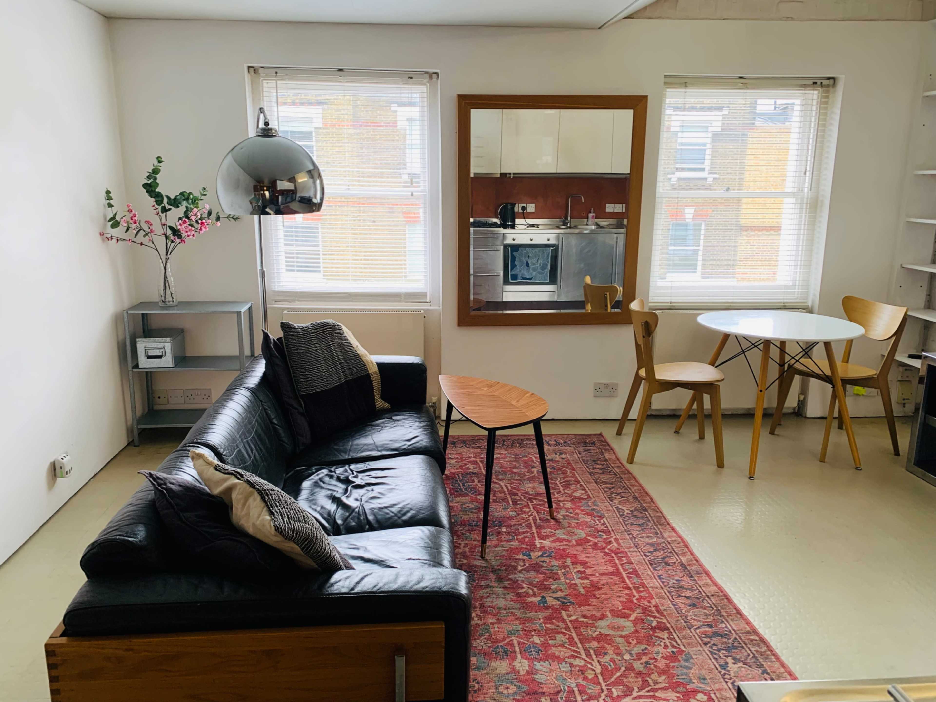 The image shows a modern living room featuring a black leather sofa, a round dining table with wooden chairs, and a large wall mirror reflecting the kitchen area.