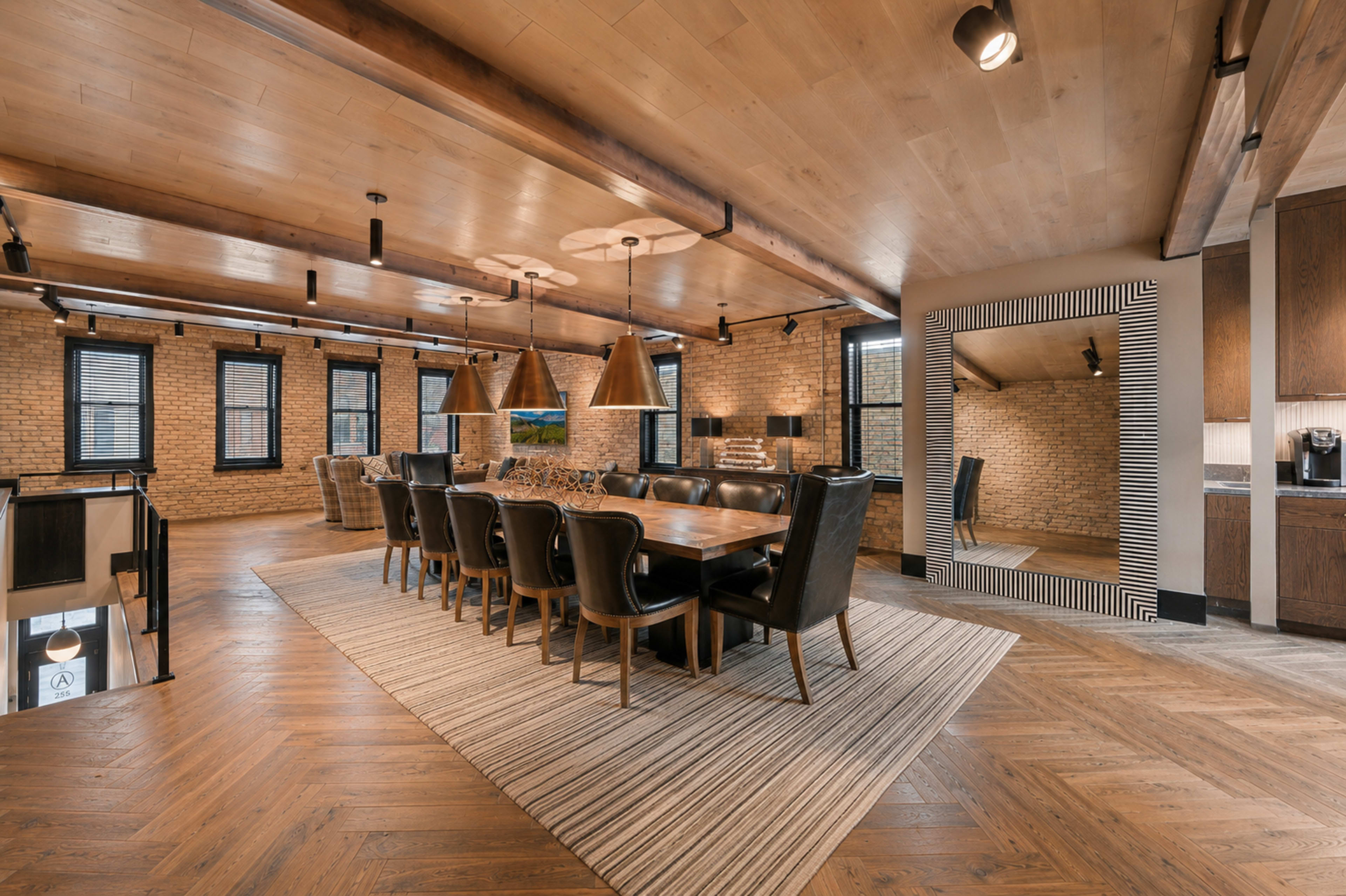 A spacious dining room features a long wooden table surrounded by black chairs, with large pendant lights above and a mirror set in a striped frame on one wall.