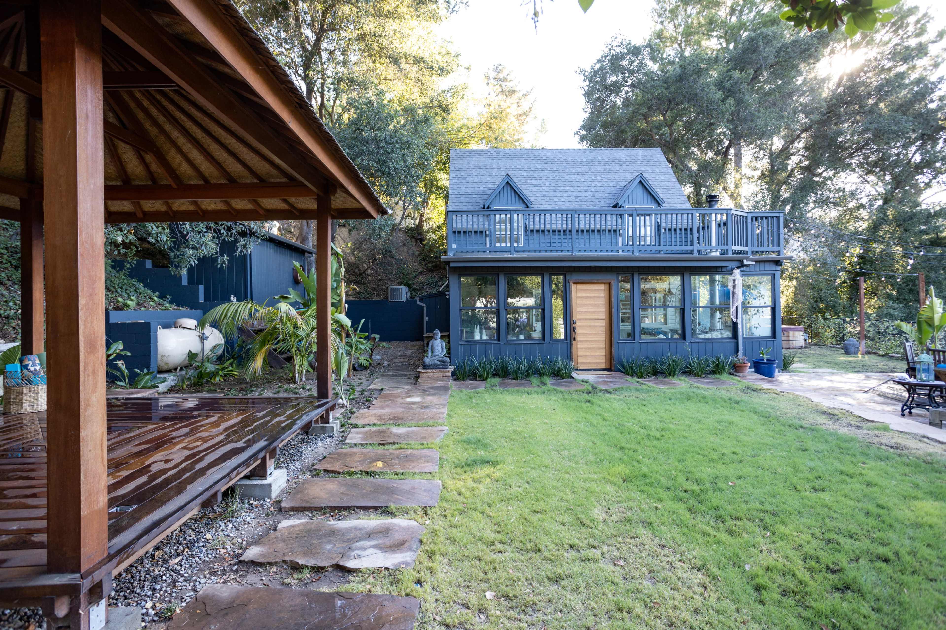 The image shows a two-story house with a peaked roof, large windows, and a wooden deck in the foreground surrounded by a neatly maintained lawn and garden.
