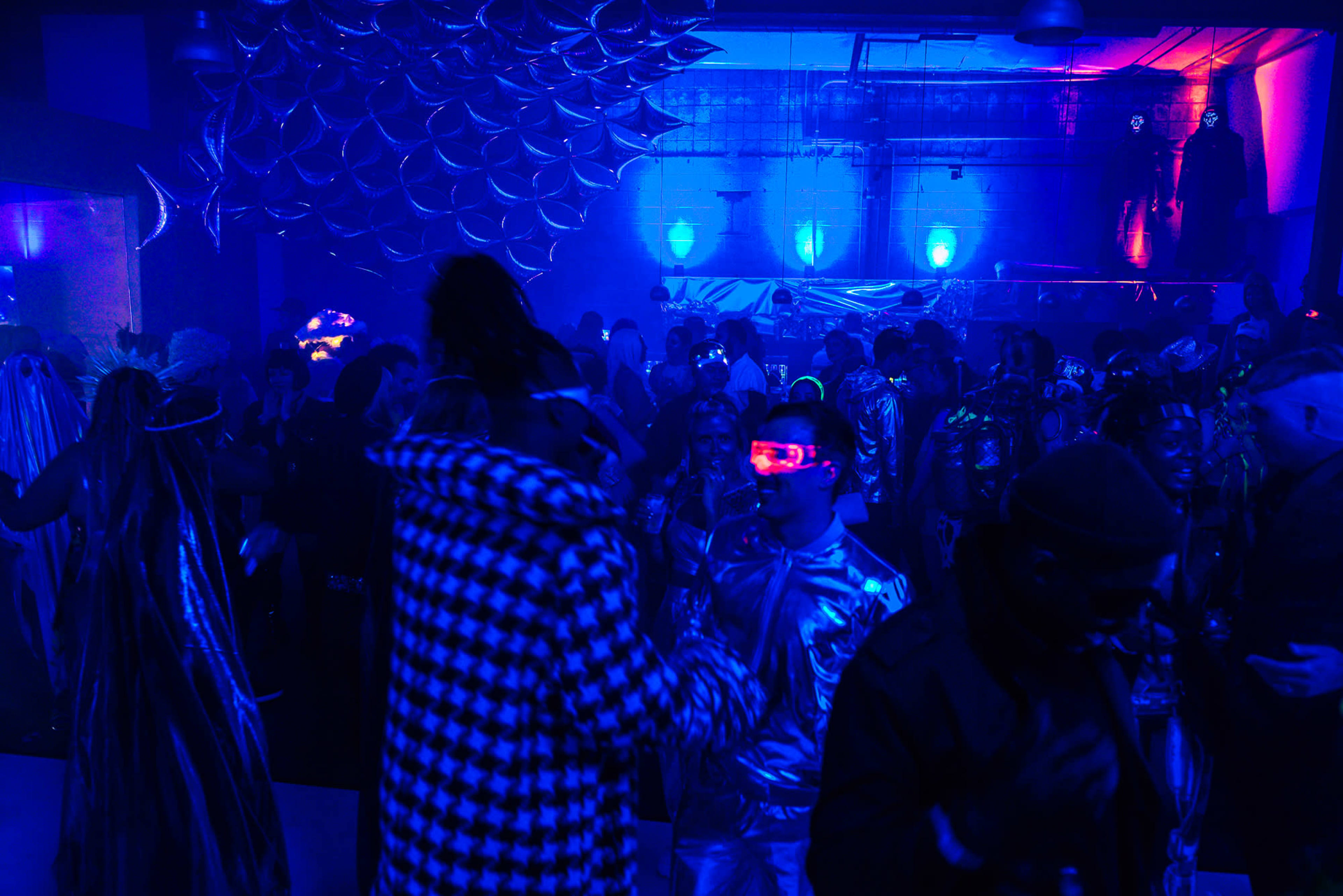 A crowd of people dressed in various costumes and reflective outfits is dancing in a dimly lit venue with blue lighting and metallic decorations.