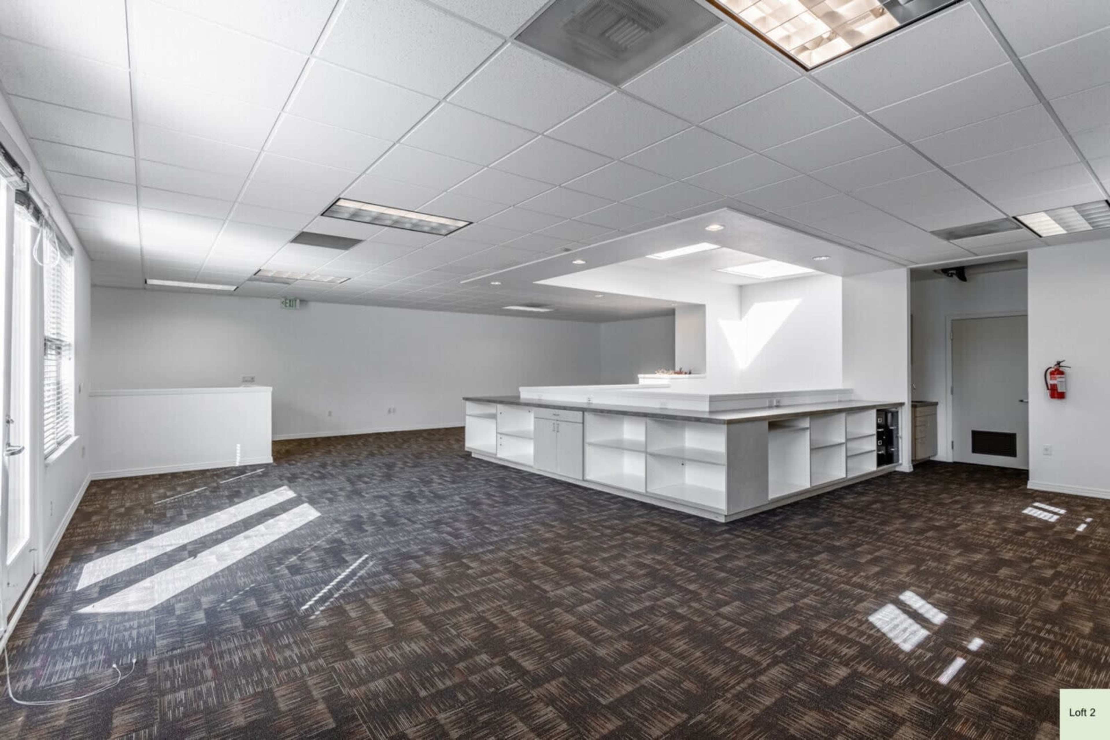 The image shows a spacious, empty office interior with a central white counter and large windows allowing natural light.