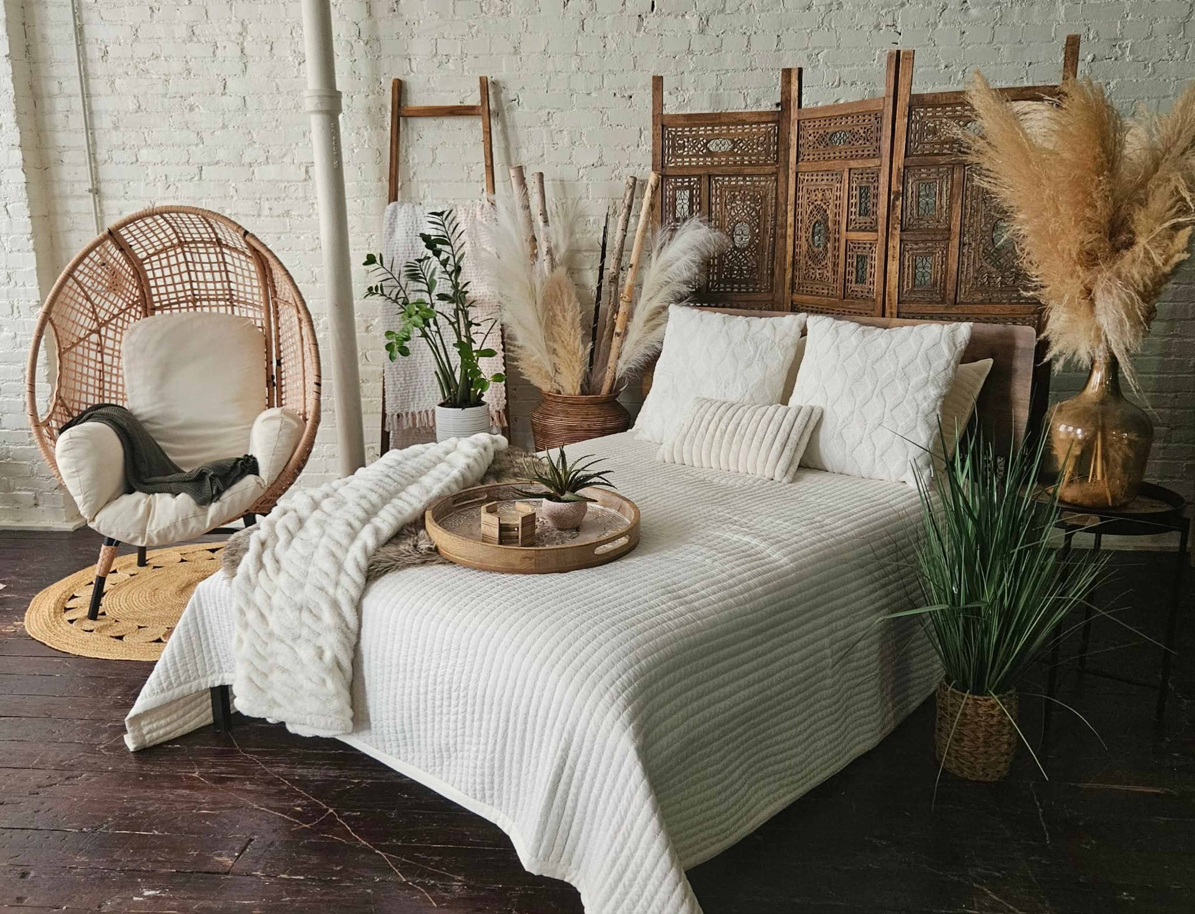 The image shows a cozy bedroom scene featuring a bed with a white quilt, a decorative wooden screen, a comfortable chair, and various plants and decorative elements arranged around the space.