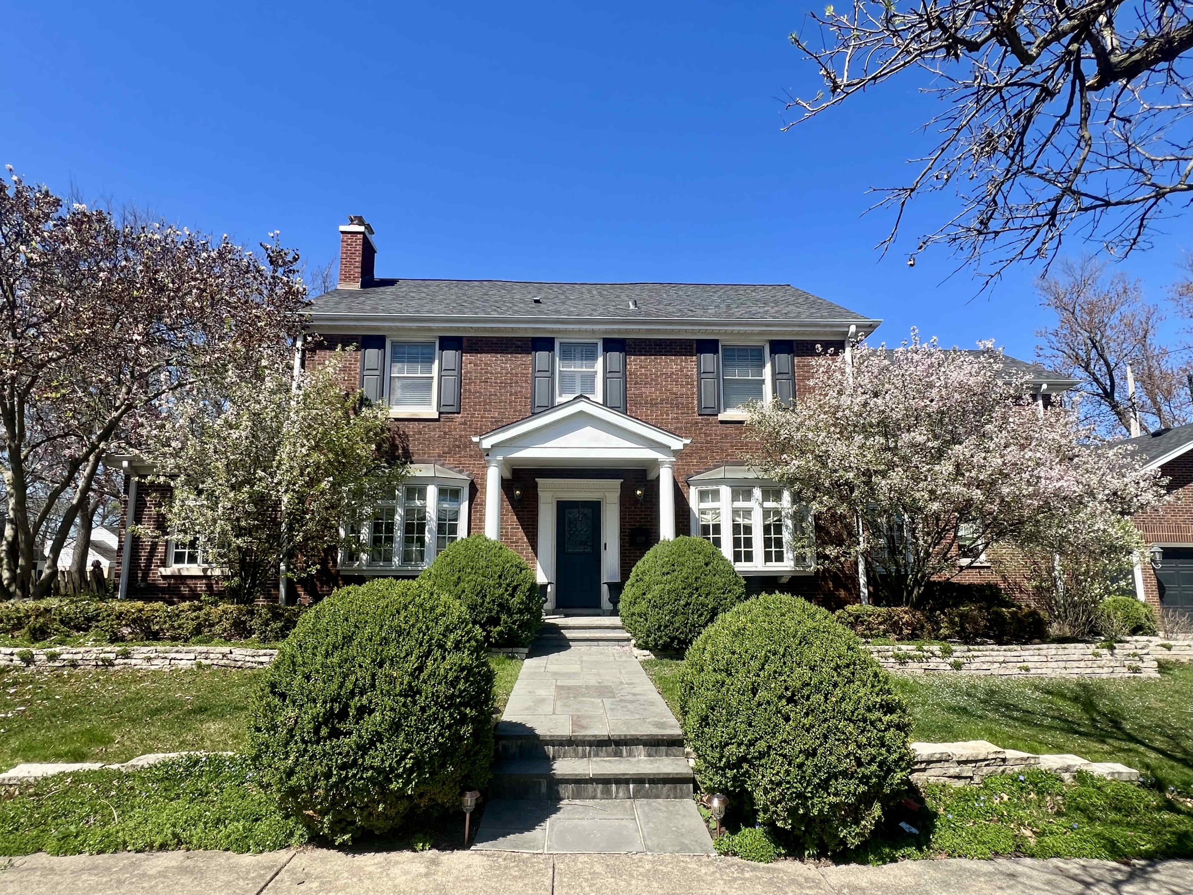 A two-story brick house with large white-framed windows, flanked by flowering trees and neatly trimmed bushes.