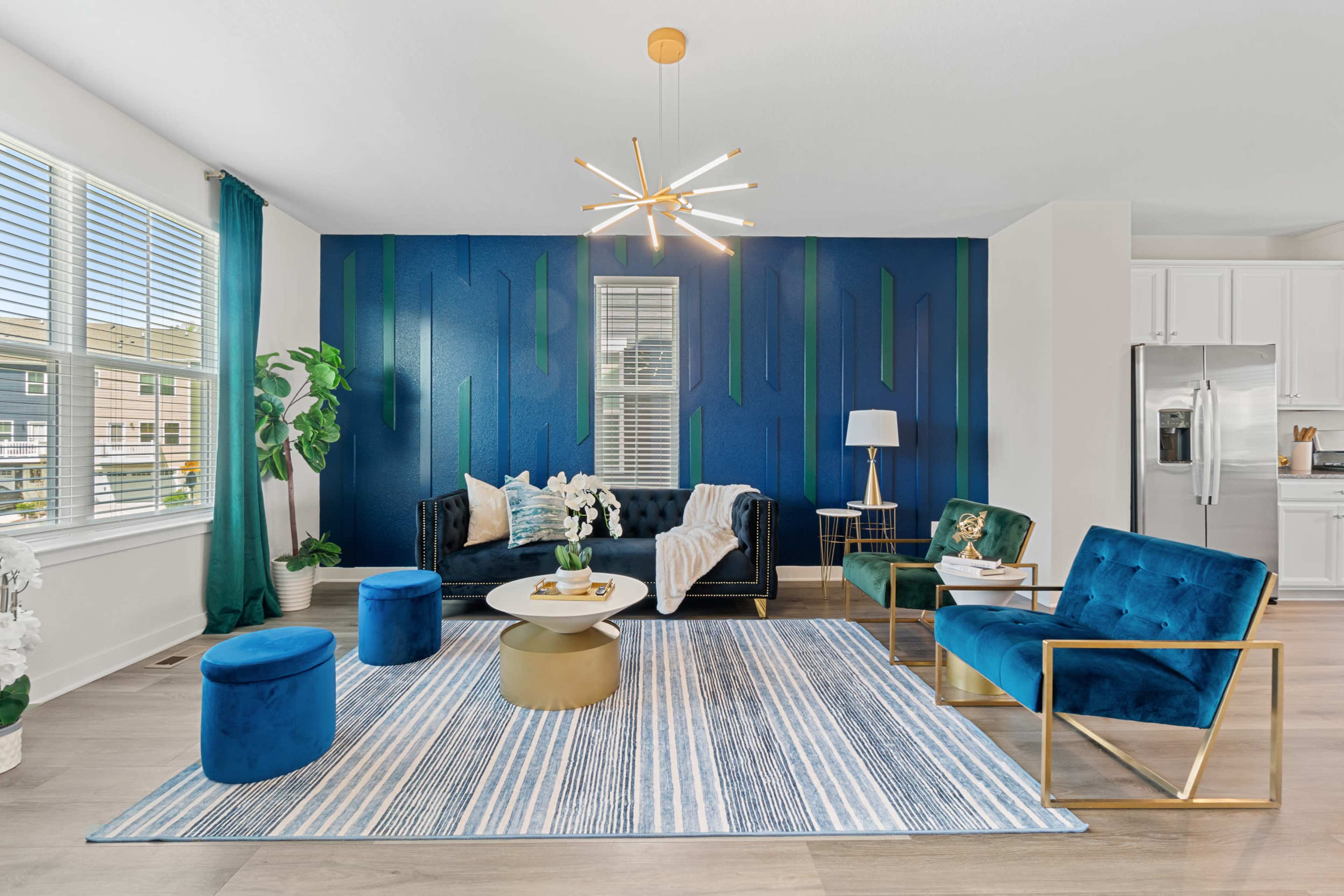 A modern living room with a dark blue accent wall, a black sofa, teal armchairs, a round coffee table, and a striped rug.