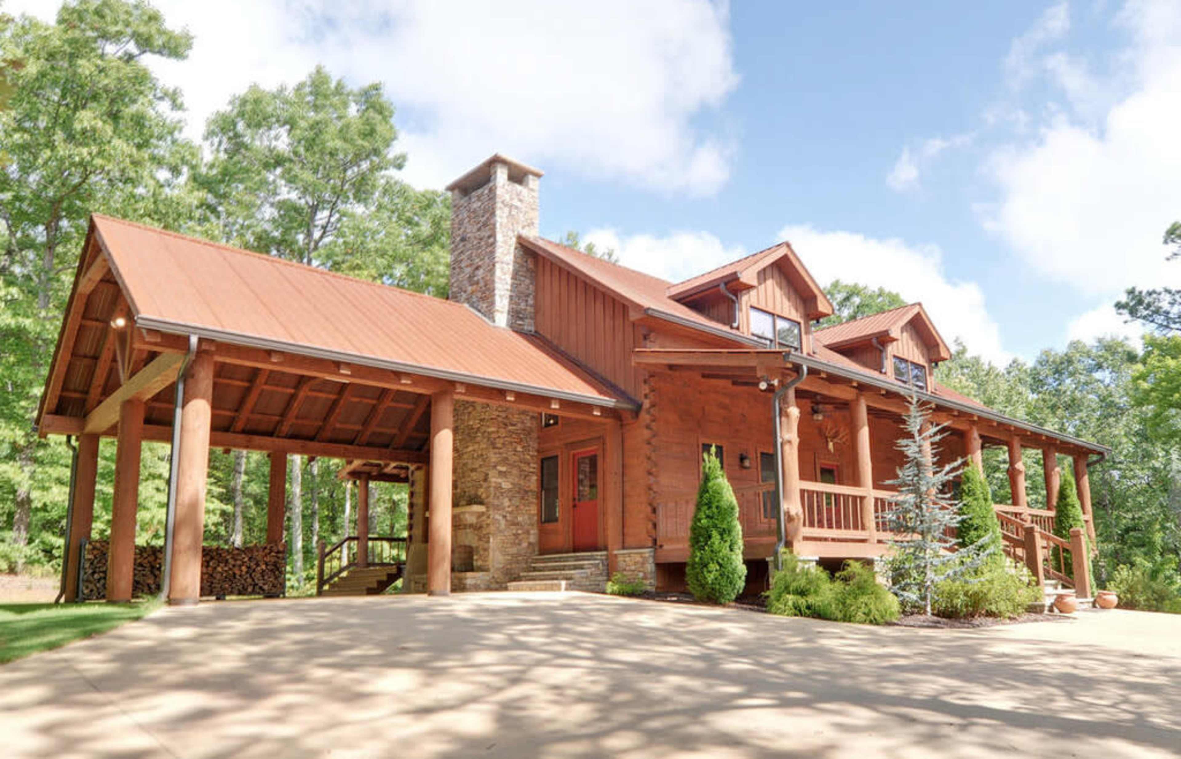 A large wooden cabin-style house with a gabled roof and a stone chimney, surrounded by trees.