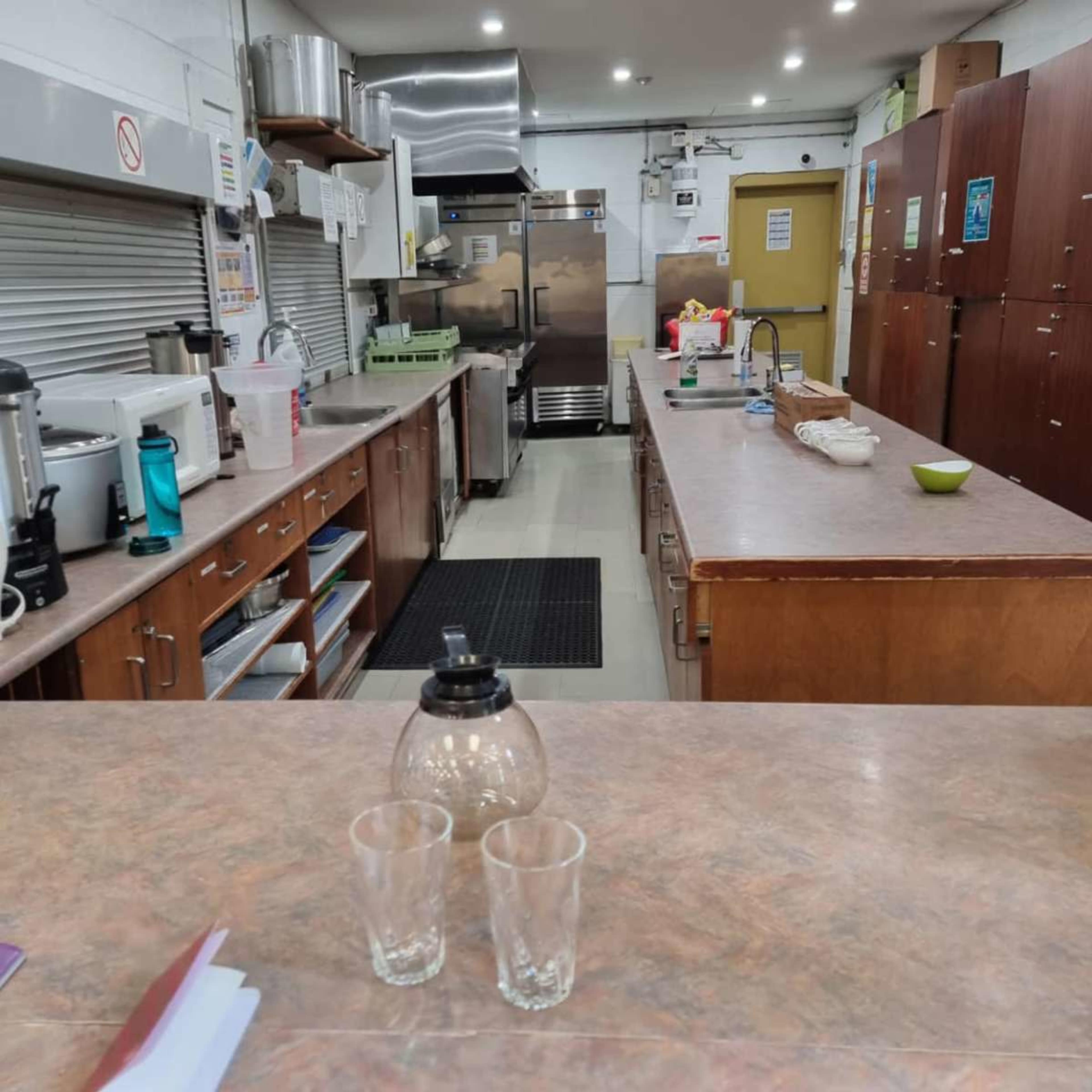The image shows a spacious commercial kitchen featuring stainless steel appliances, wooden cabinets, and a long countertop with two glass cups.