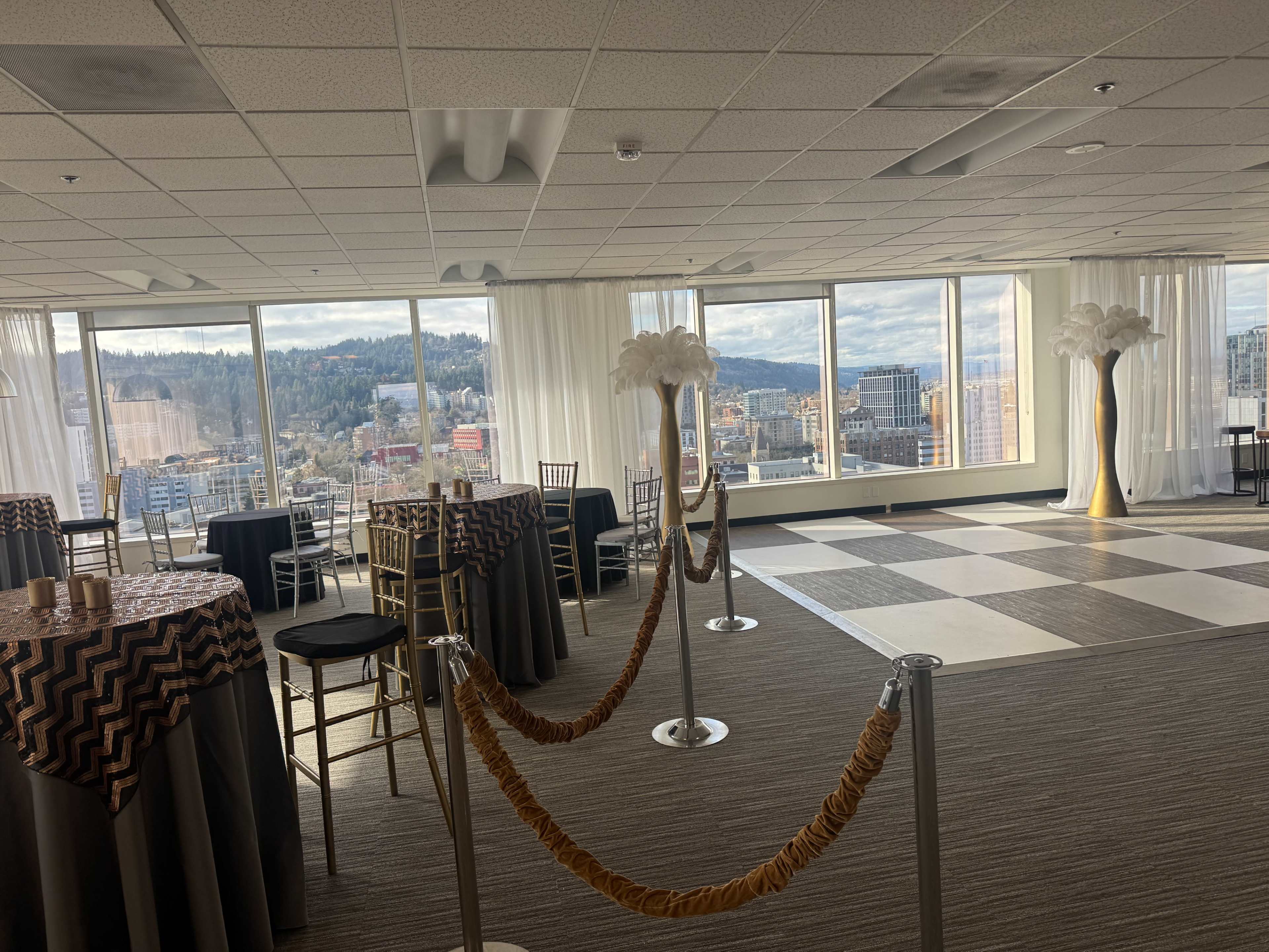 The image shows a spacious event room with large windows overlooking a cityscape, featuring elegantly dressed tables and decorative lighting.