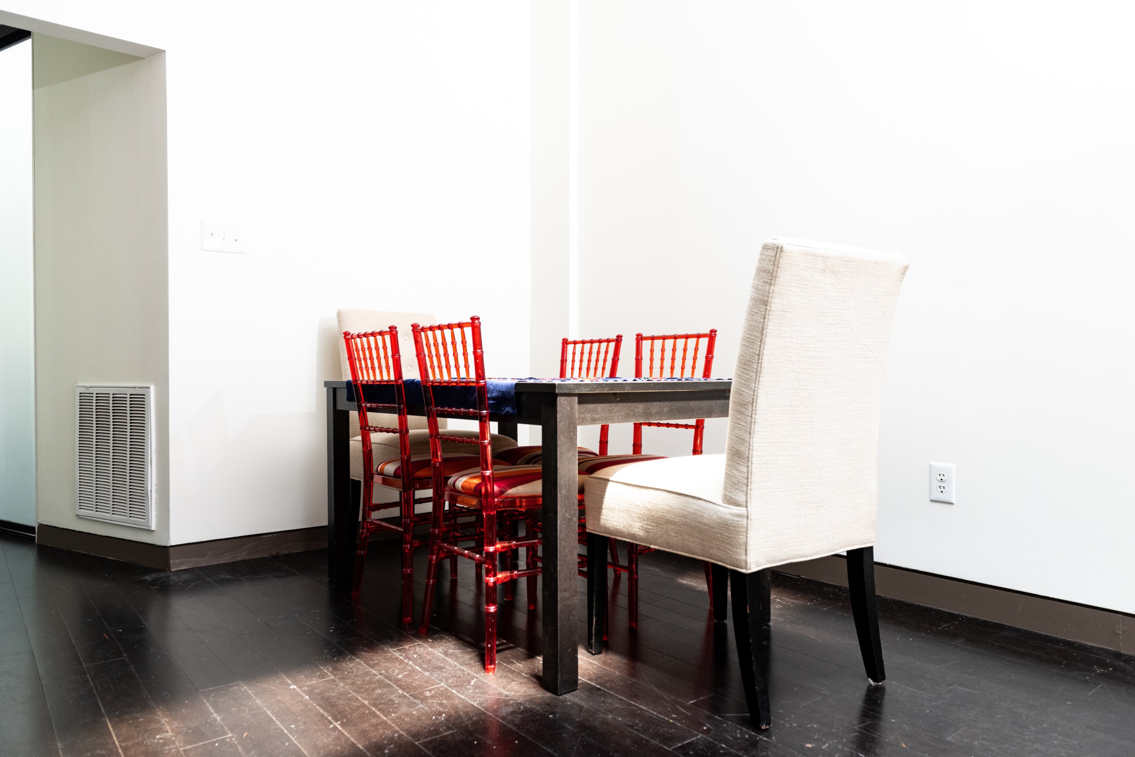 A wooden table surrounded by red chairs and a single beige upholstered chair is positioned in a well-lit room with light-colored walls.