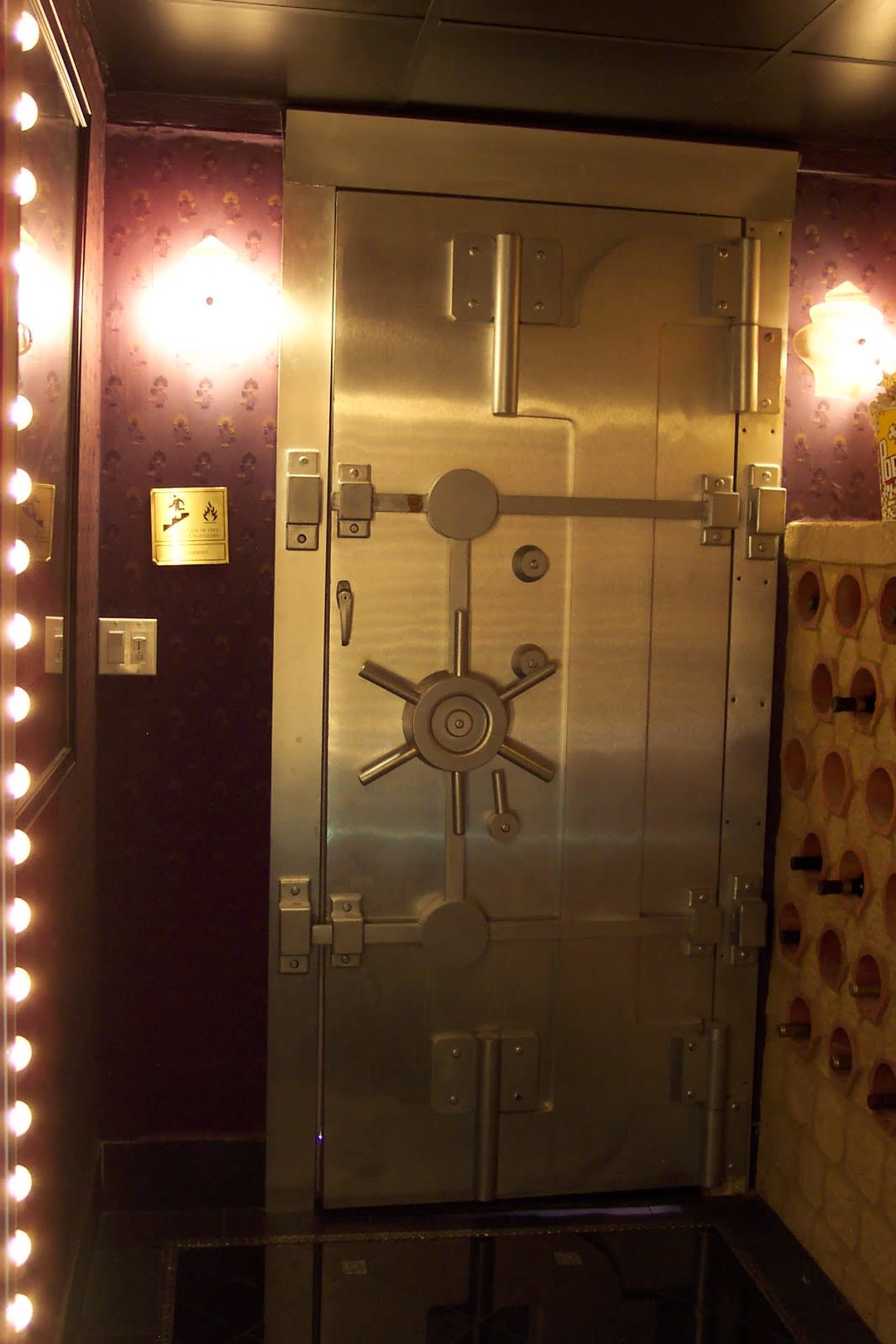 A large, metallic vault door with a wheel mechanism, set against a wall featuring purple wallpaper and bright lights.