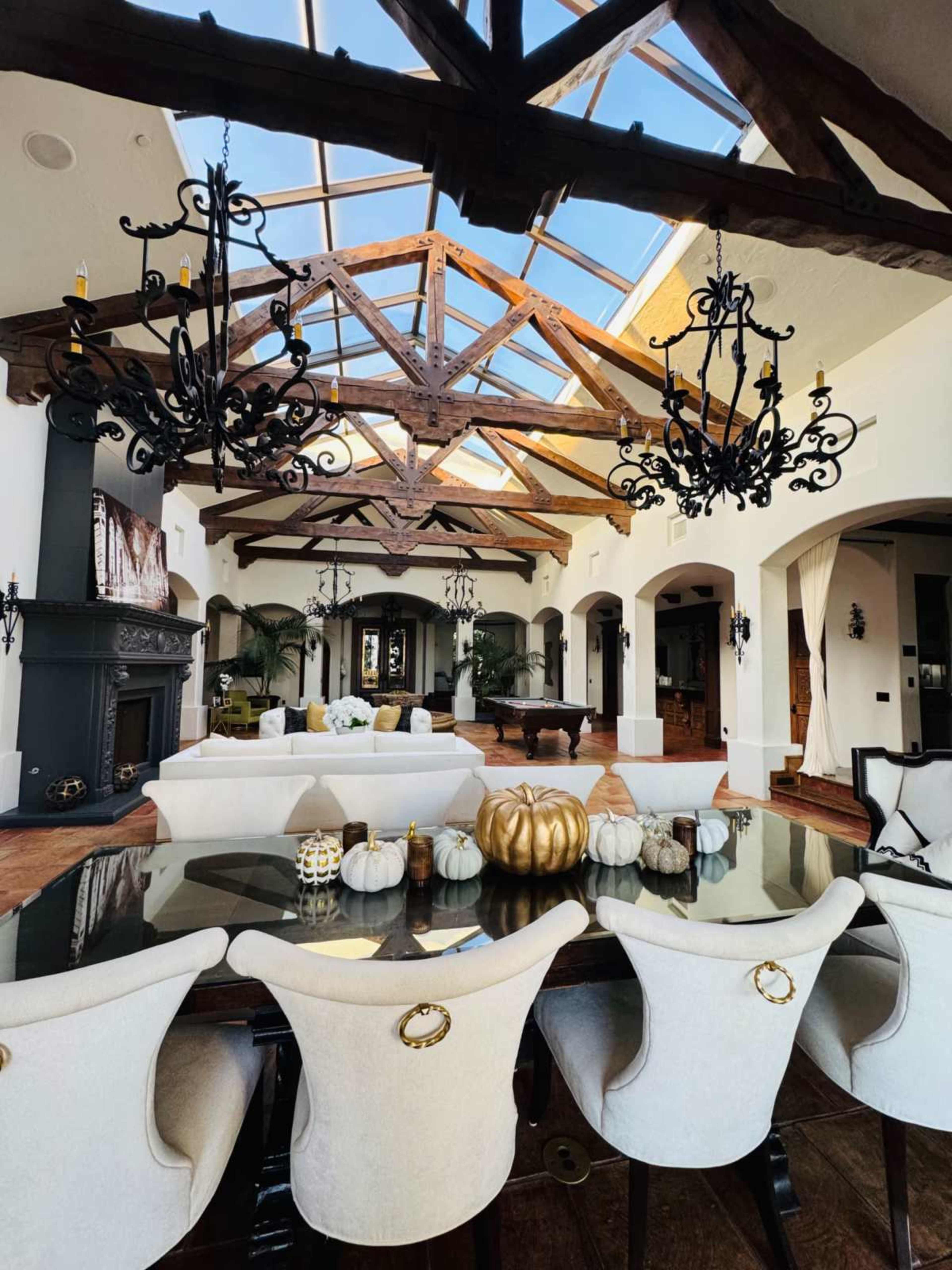 The image shows an elegantly designed living space with a large wooden dining table, plush chairs, and a high ceiling featuring exposed beams and skylights.