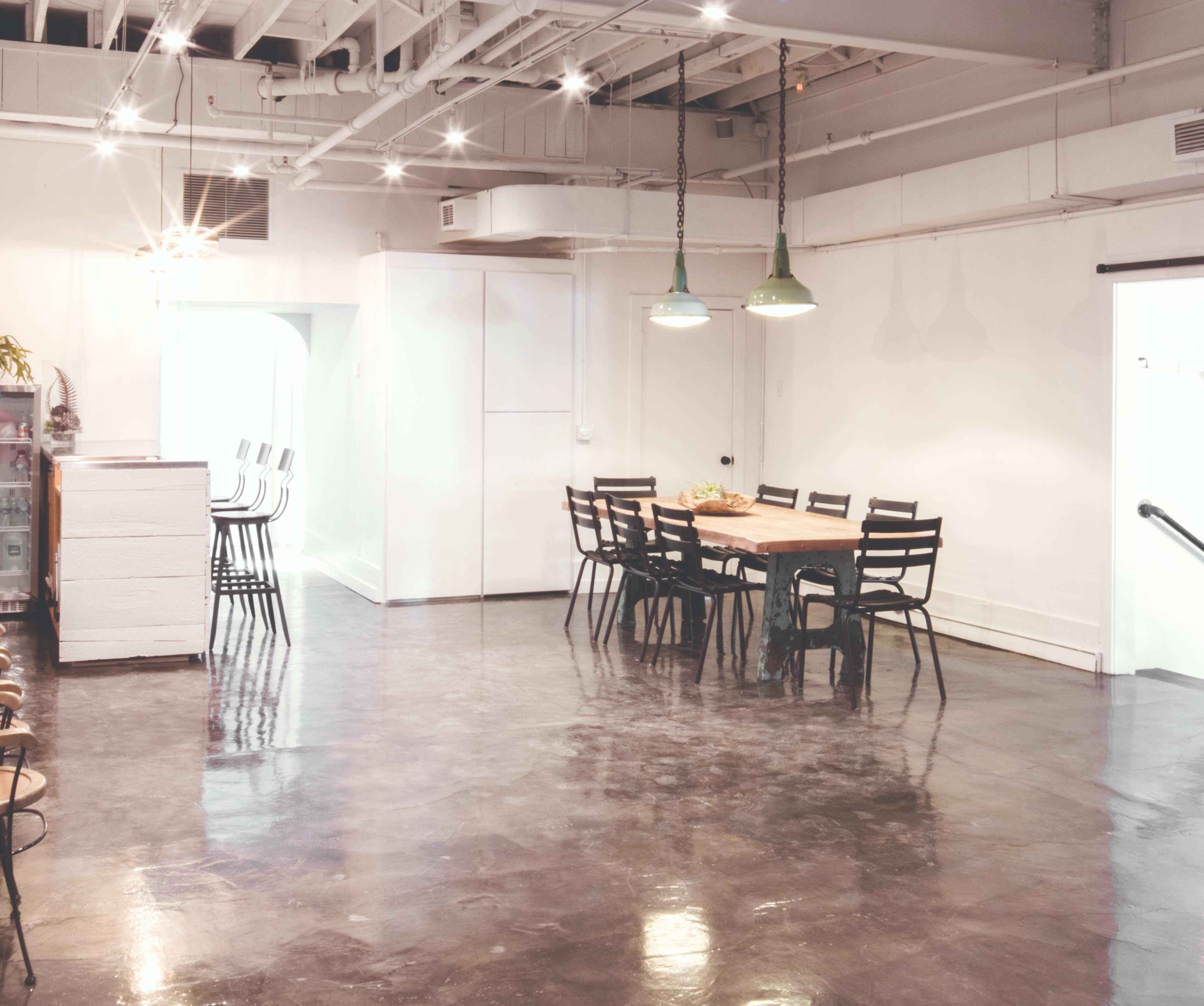 A spacious, brightly lit room features a large wooden dining table surrounded by black chairs, with a polished concrete floor and white walls.