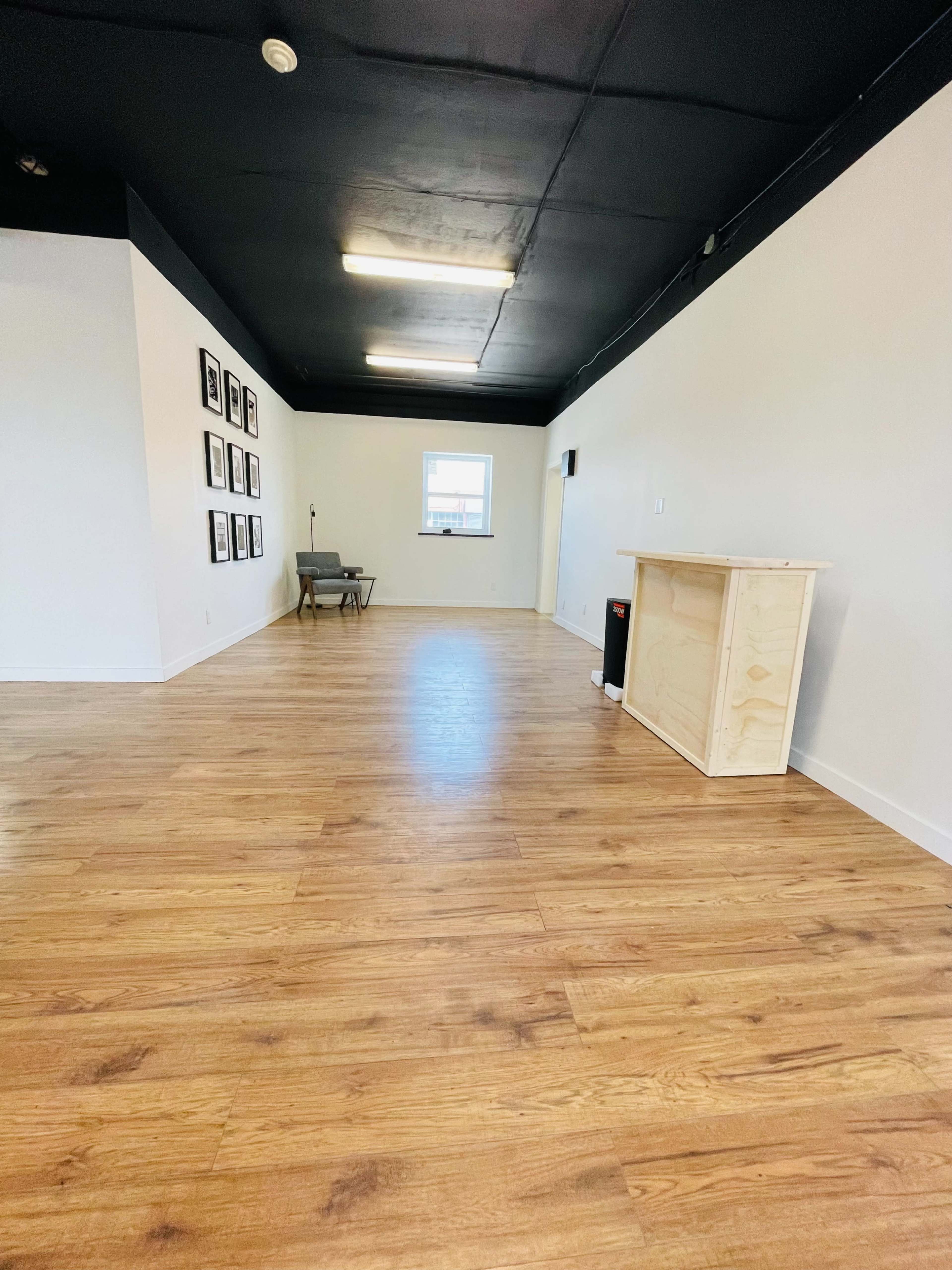 A spacious, empty room with wooden flooring, a wall of picture frames on one side, and a small wooden counter placed near the entrance.