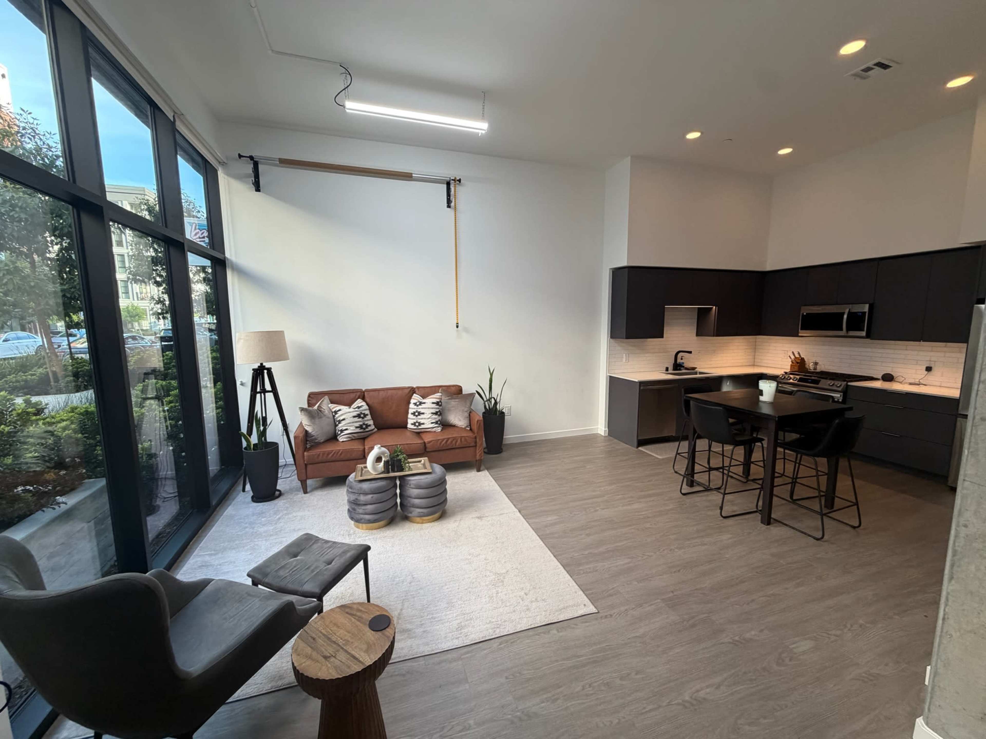 The image shows a modern living space featuring a brown sofa with decorative pillows, a dining area with black chairs and a kitchen, and large windows letting in natural light.