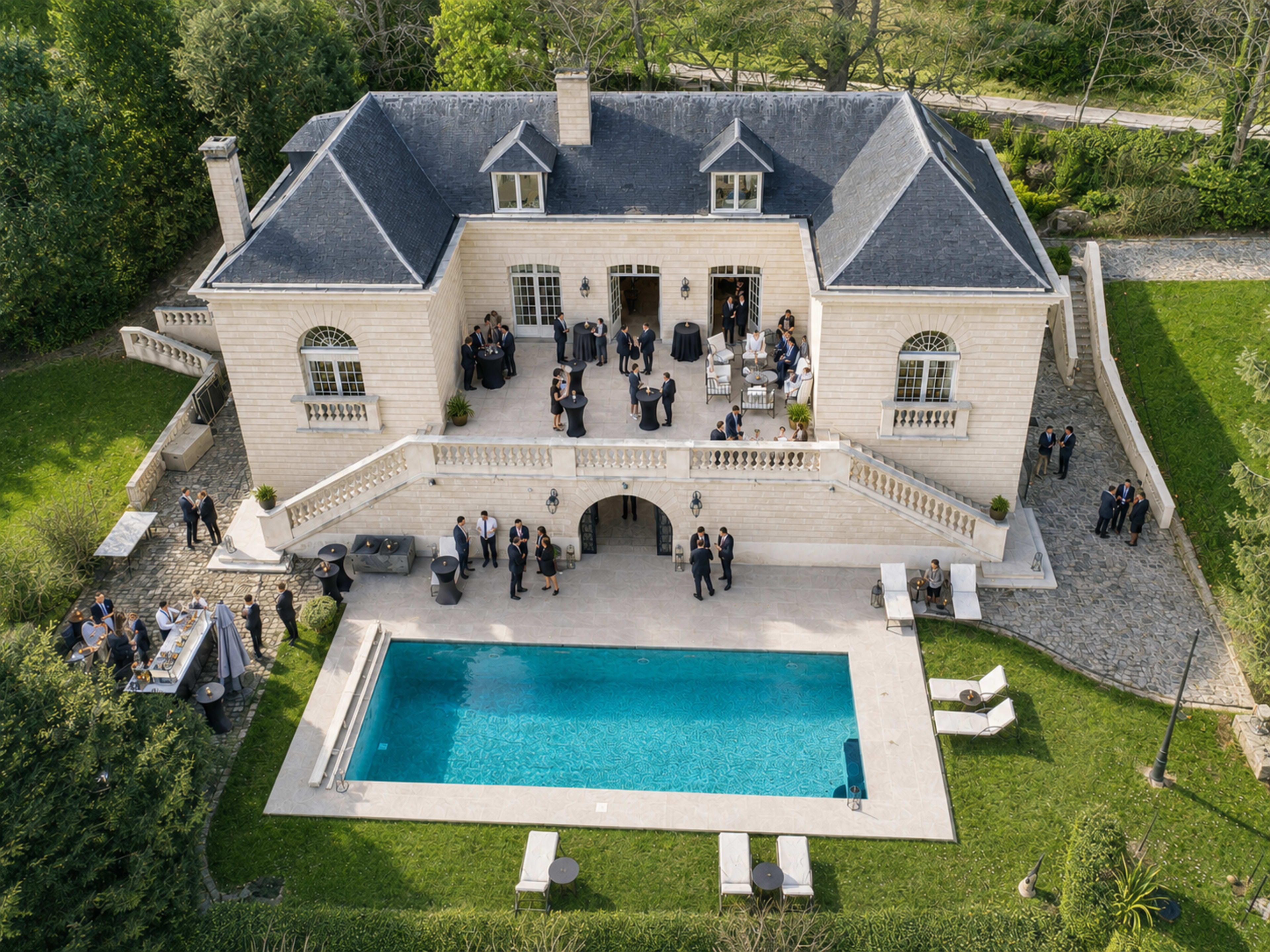 An aerial view shows a large, elegant mansion with a swimming pool and a gathering of people on the terrace and surrounding areas.