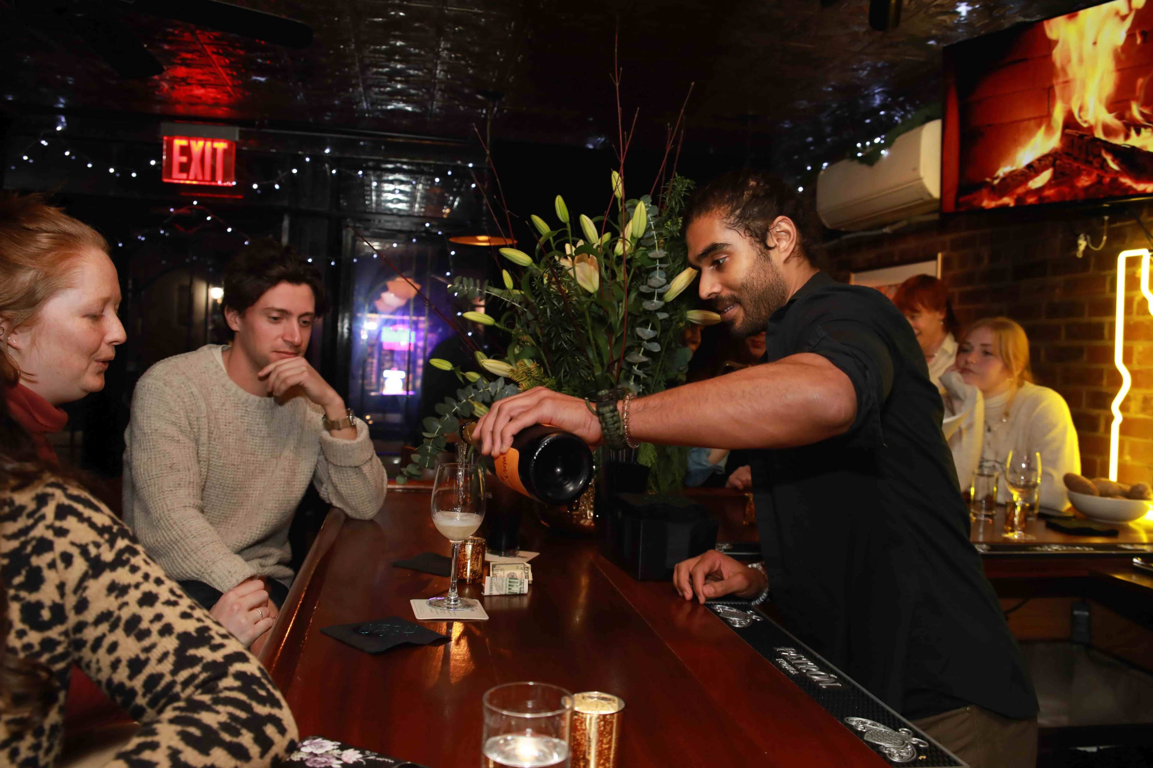 A bartender pours a drink for two seated customers at a warmly lit bar adorned with floral arrangements.