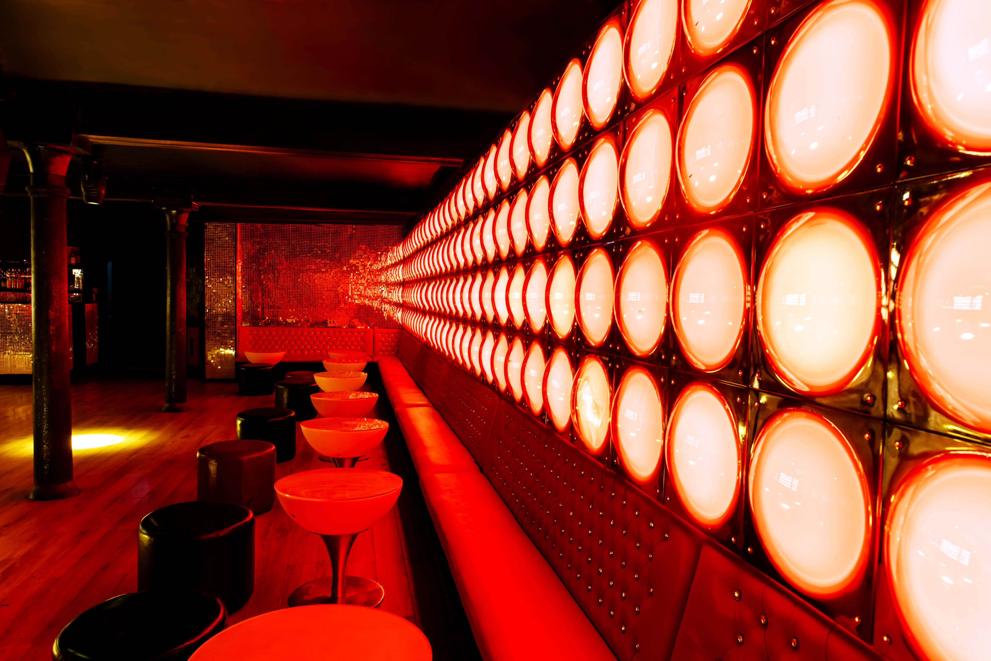 The interior of a bar features a wall of round, illuminated panels with a warm red glow, surrounded by a mix of seating options.