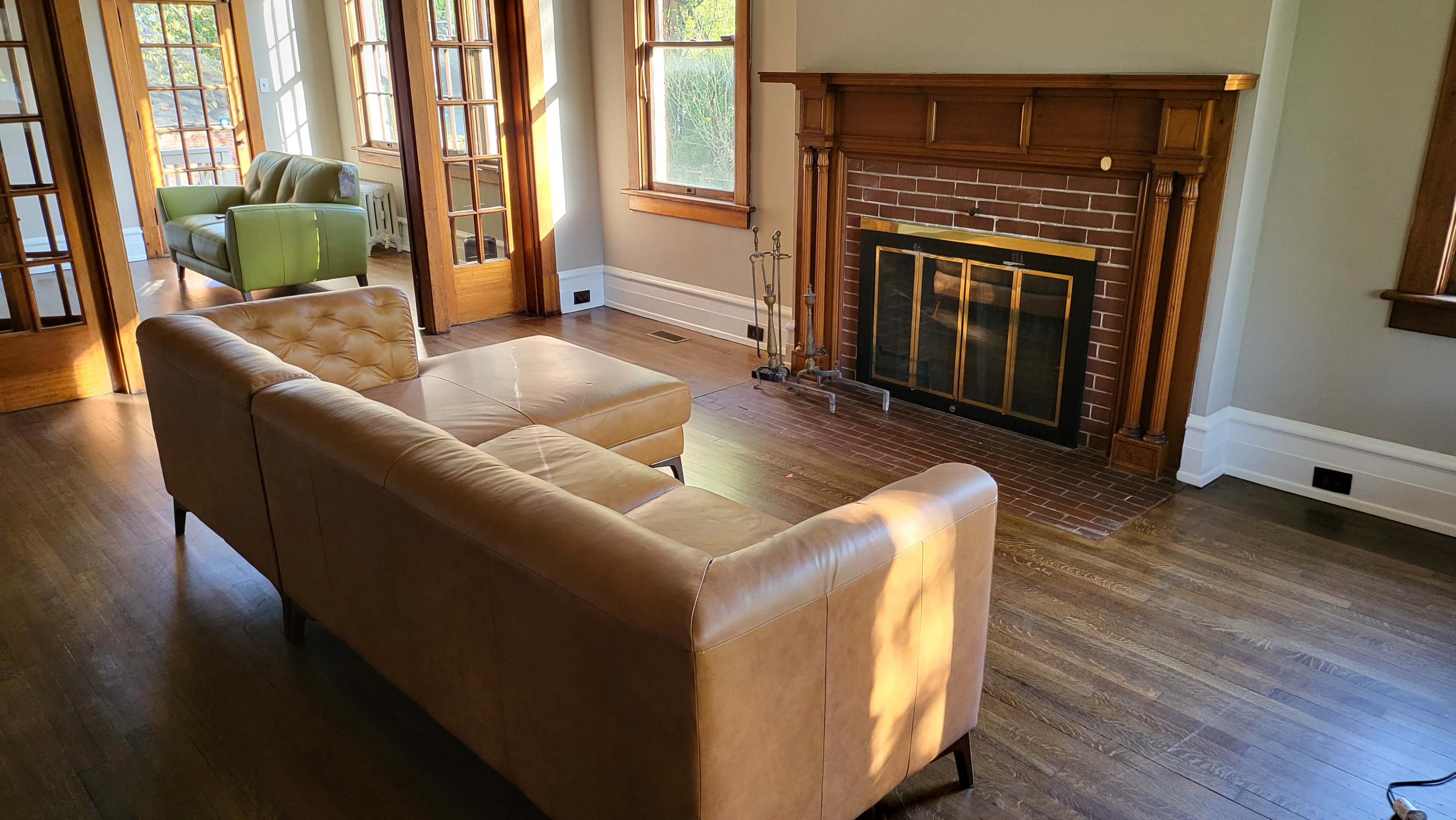A living room featuring a tan leather sofa, a brick fireplace, and large windows that allow natural light to enter the space.