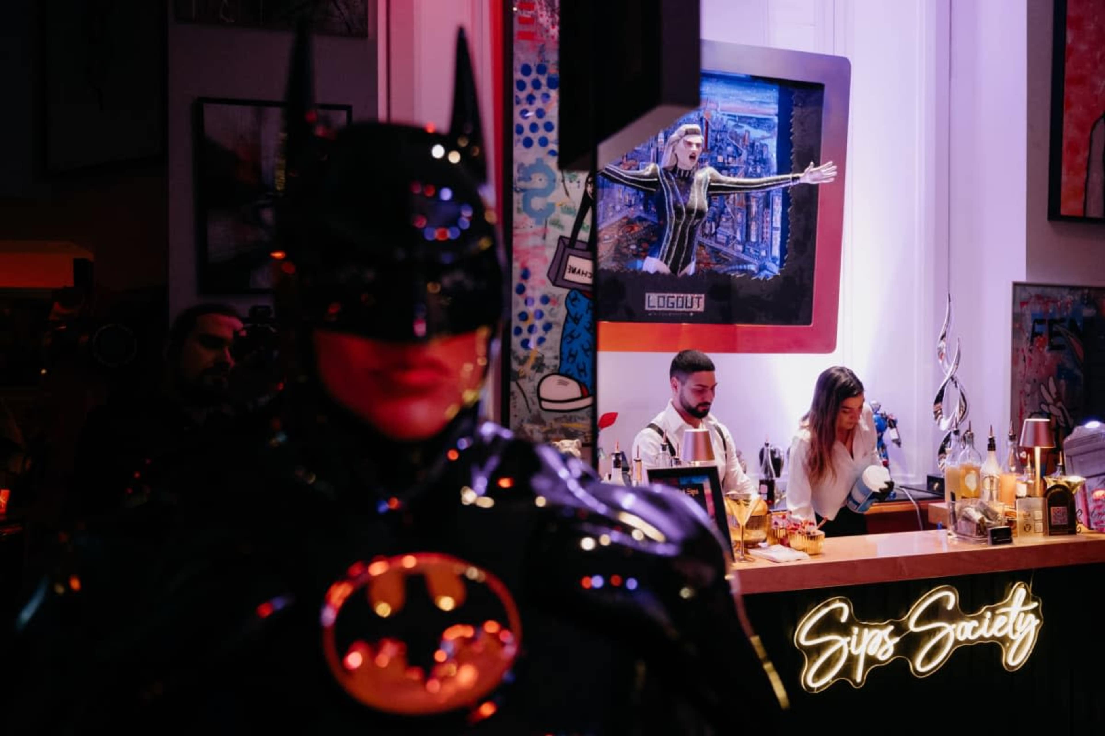 The image features a bar scene with a large Batman figurine in the foreground and a bartender serving drinks in the background, alongside a woman at a counter illuminated by colorful lights.