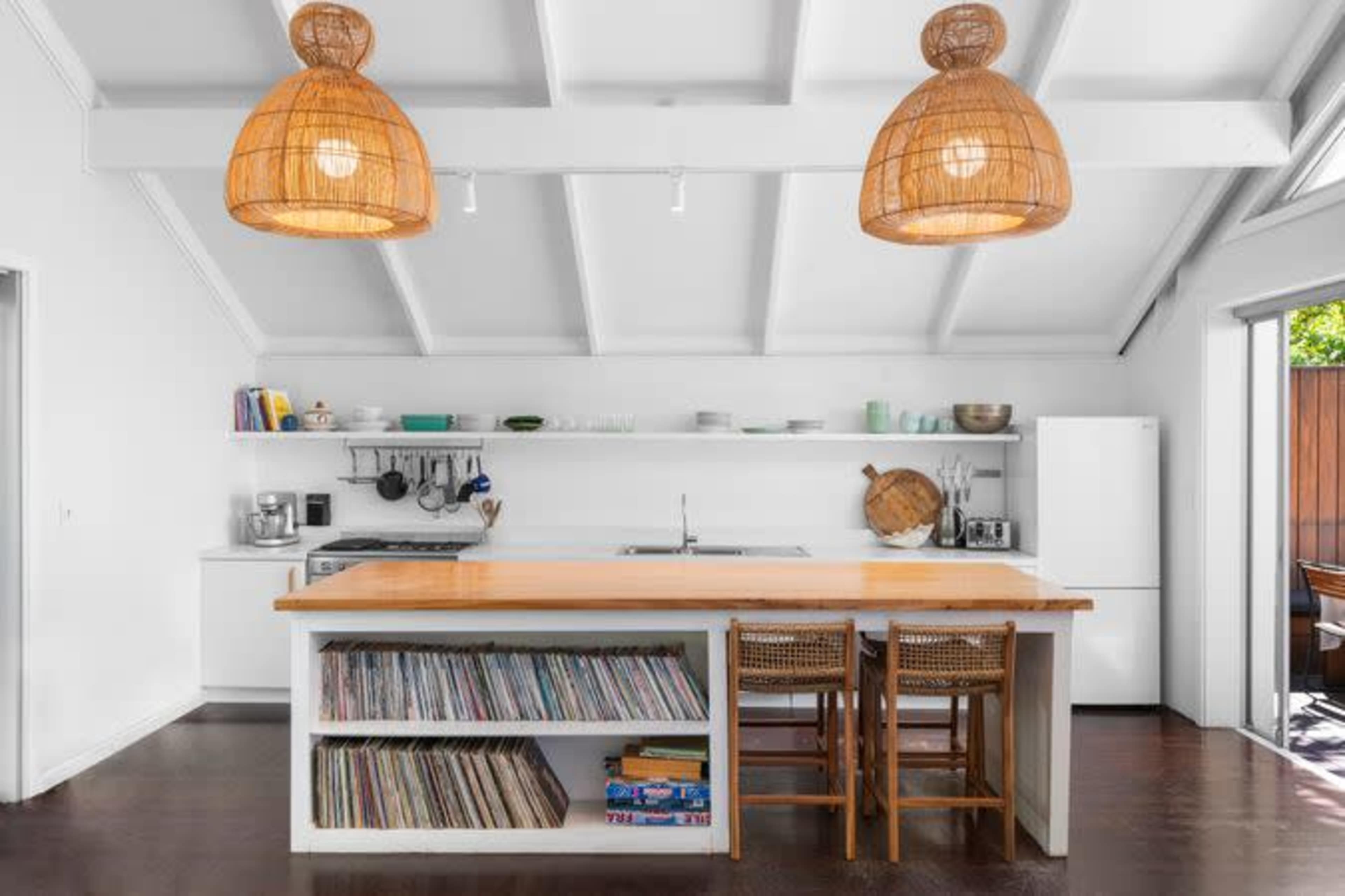 A modern kitchen features white walls, a wooden island with chairs, and two woven pendant lights hanging from the ceiling.
