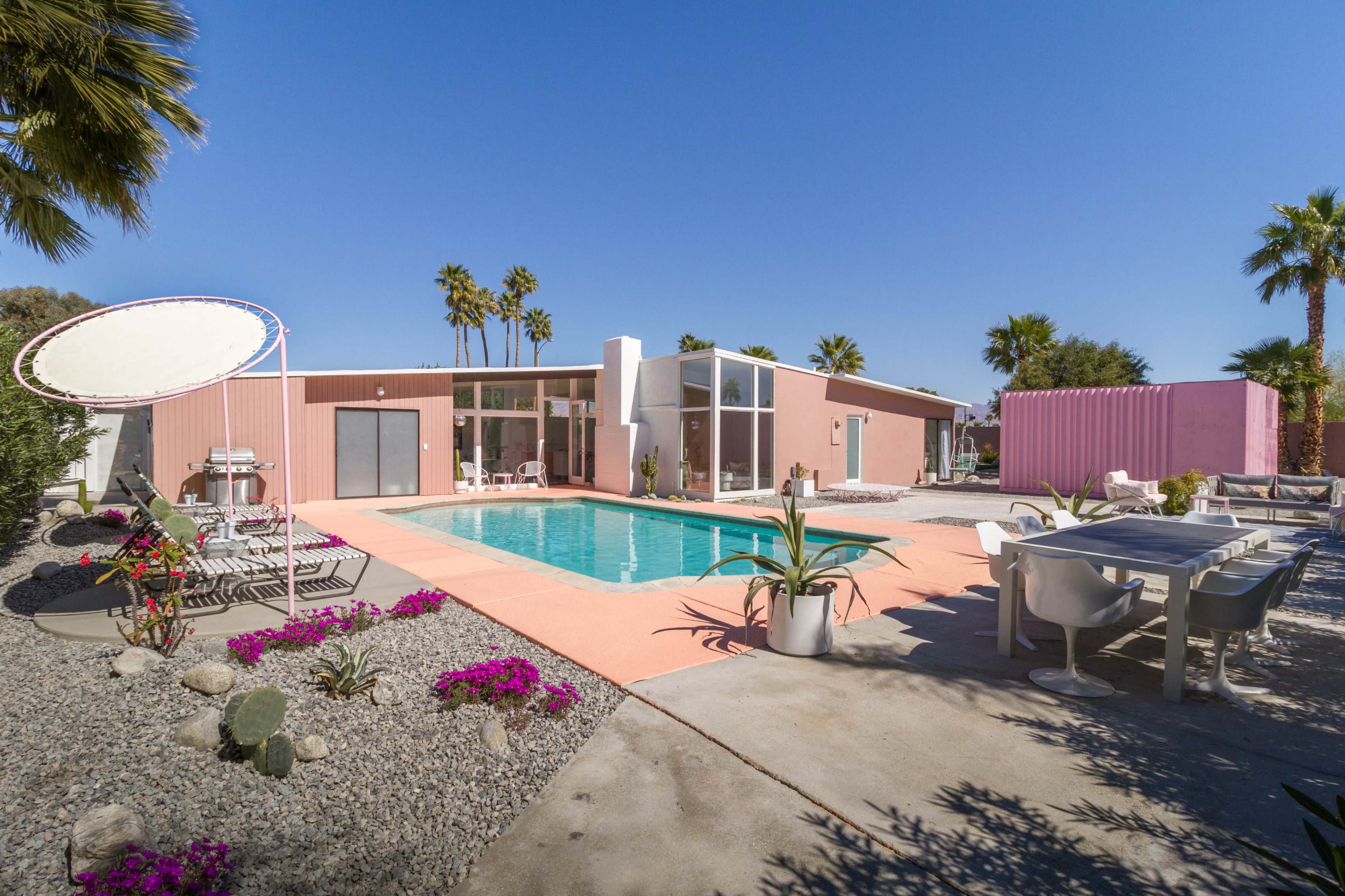 A modern pink and white house with a swimming pool surrounded by palm trees and landscaped gravel areas.